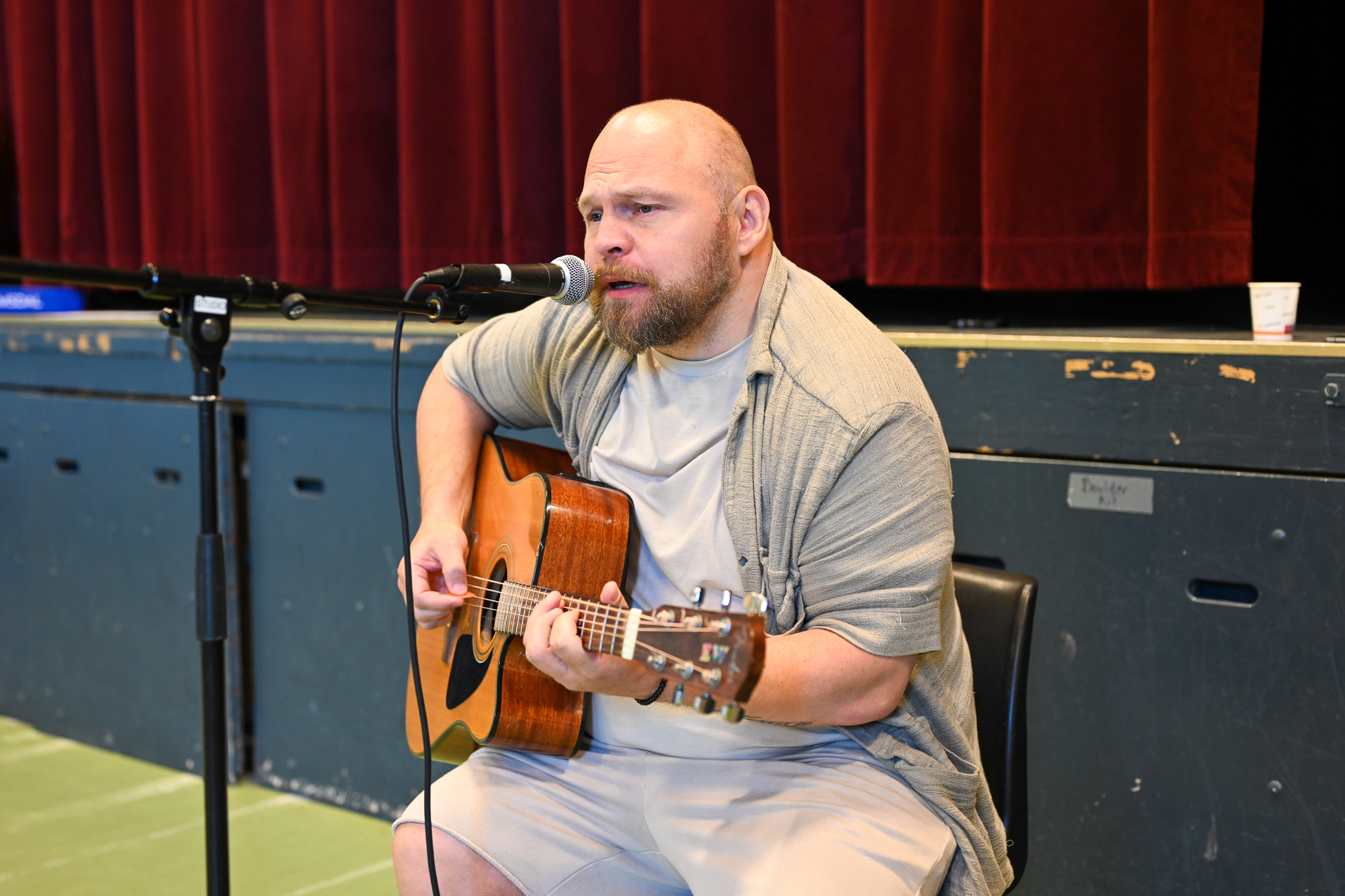 Mann med gitar synger i mikrofonen. Han sitter i en gymsal.