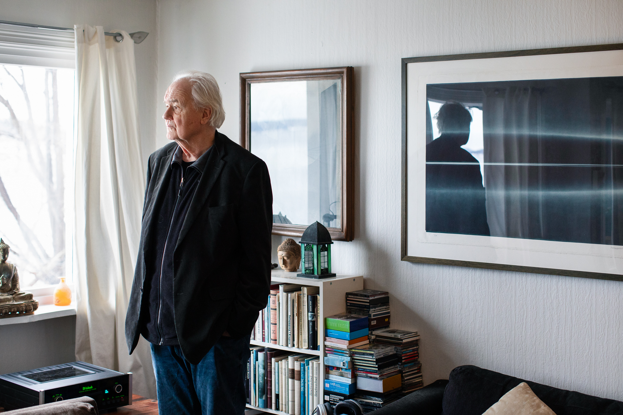 Eldre mann i dressjakke og dongeribukse står i en stue. På veggen henger kunst, i bokhyllene bak ham står mange bøker. Han ser ut mot et vindu.