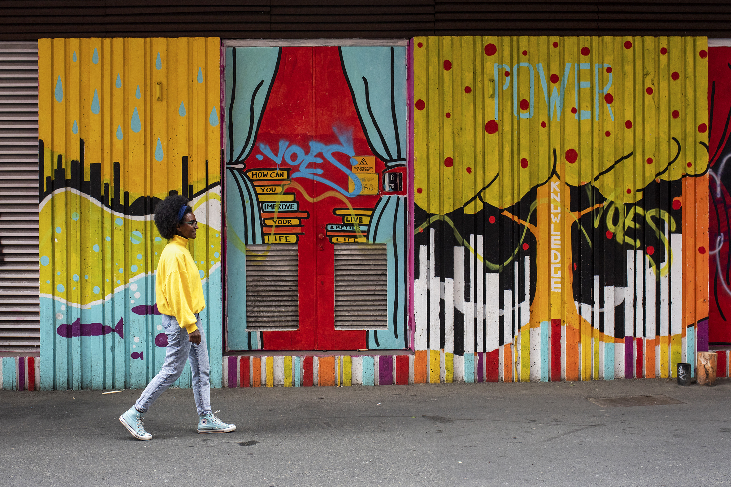 Kidist går forbi en vegg malt i sterke farger, med blant annet trær og vann med fisker malt. Hun har på seg knallgul genser og jeans,