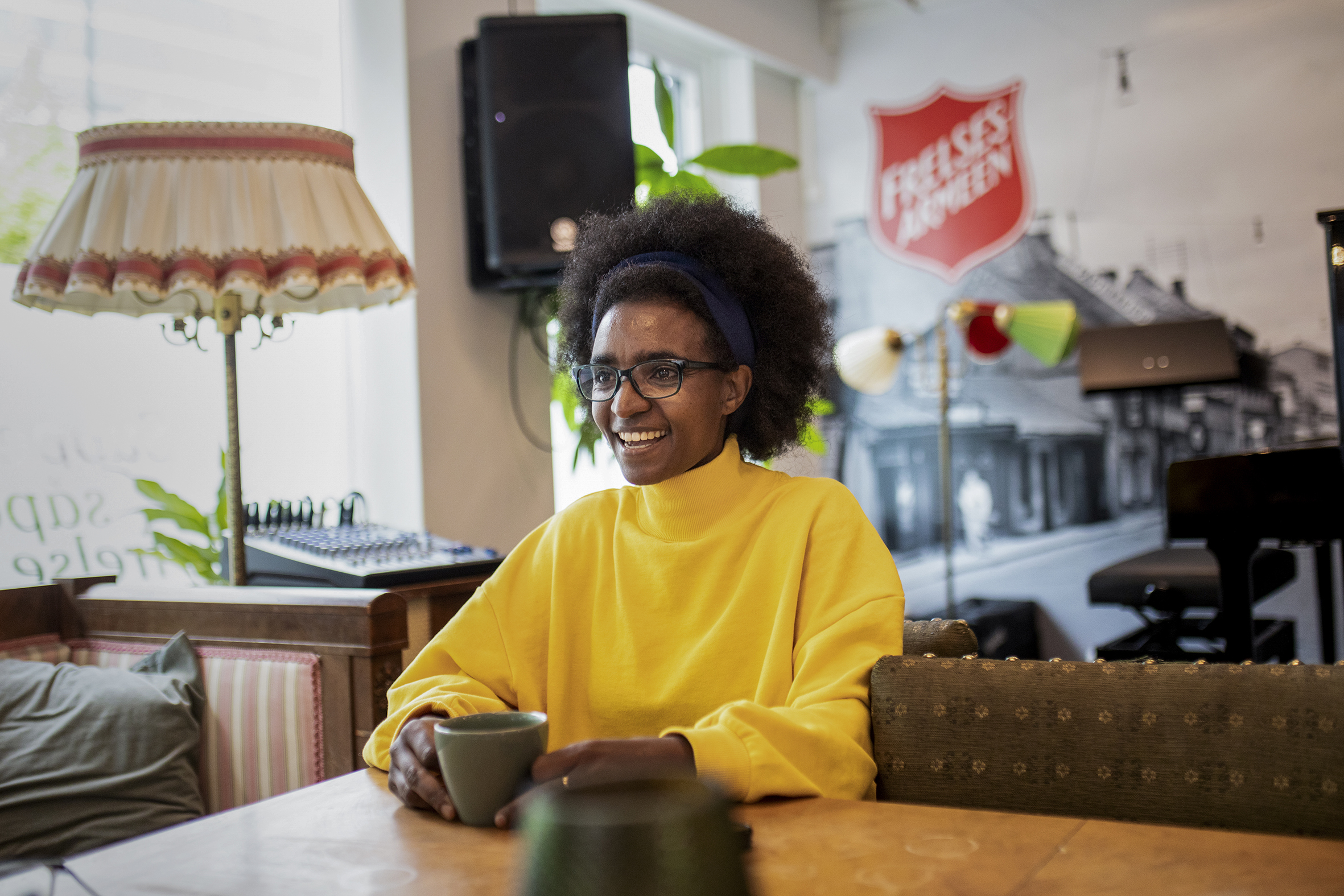 Kidist sitter i kafeen på Sammen. Bak henne er et stort svart-hvitt bilde fra Oslo før i tiden, og møbler og en lampe i retro-stil.