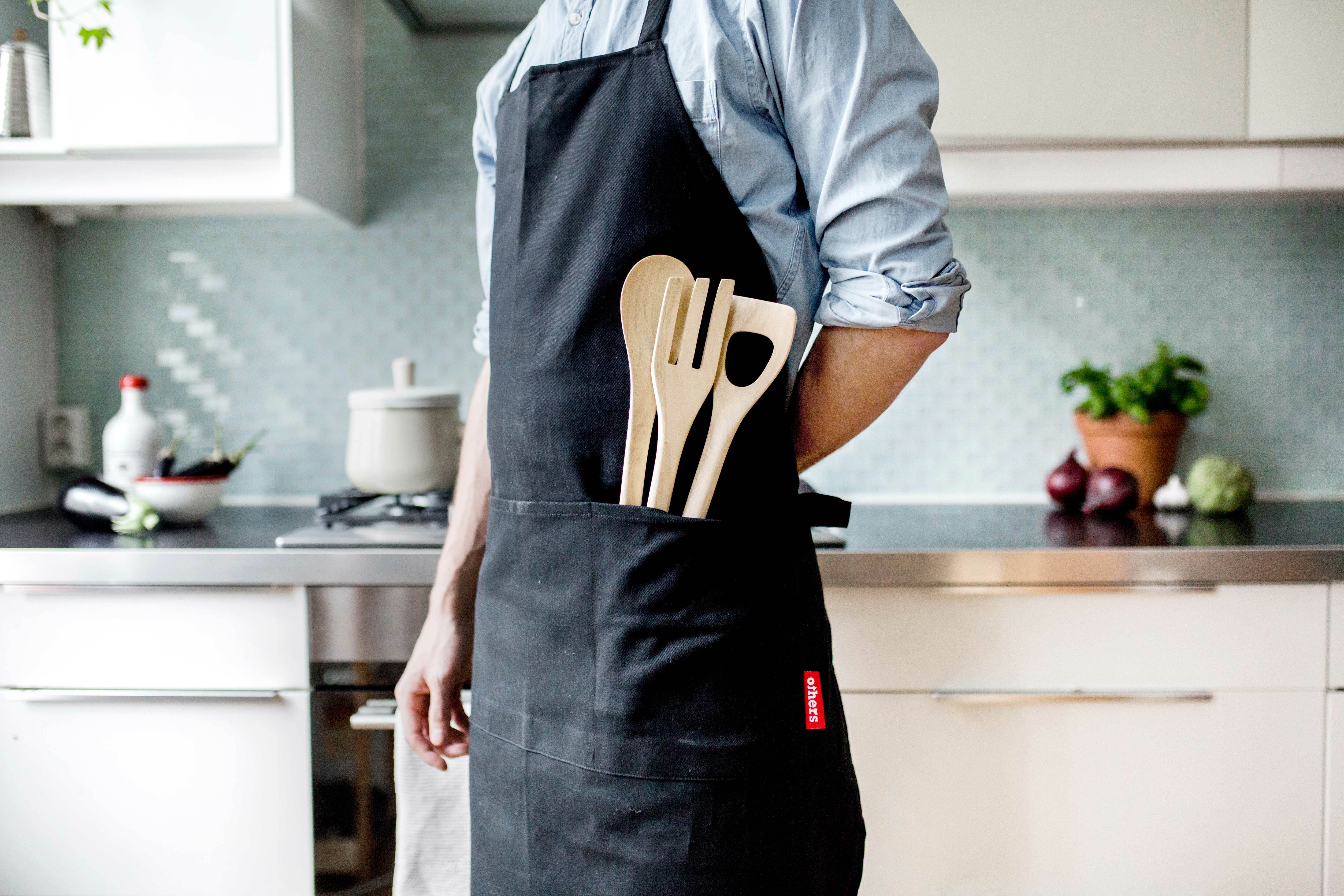 Mann står med forkle på et kjøkken. I den ene lommen stikker det opp kjøkkenredskaper av tre.