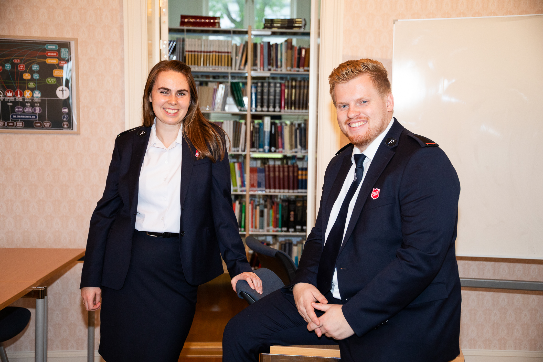 Ungt par i uniform smiler mot kamera i et rom med rosa tapet på veggene og bøker i rommet bak.