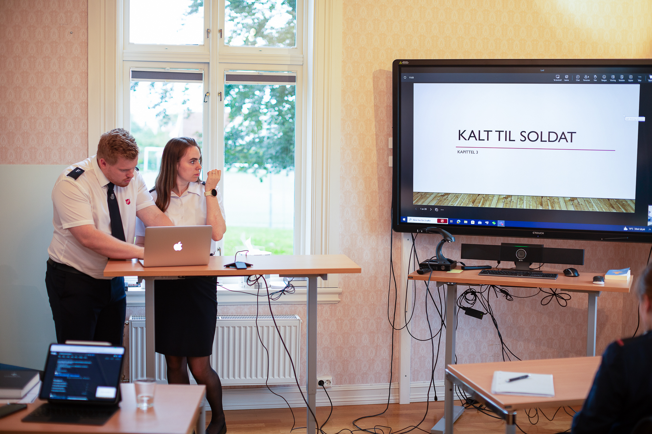 Par i uniform står bak en PC i et klasserom. Har fått tekst opp på en TV-skjerm.