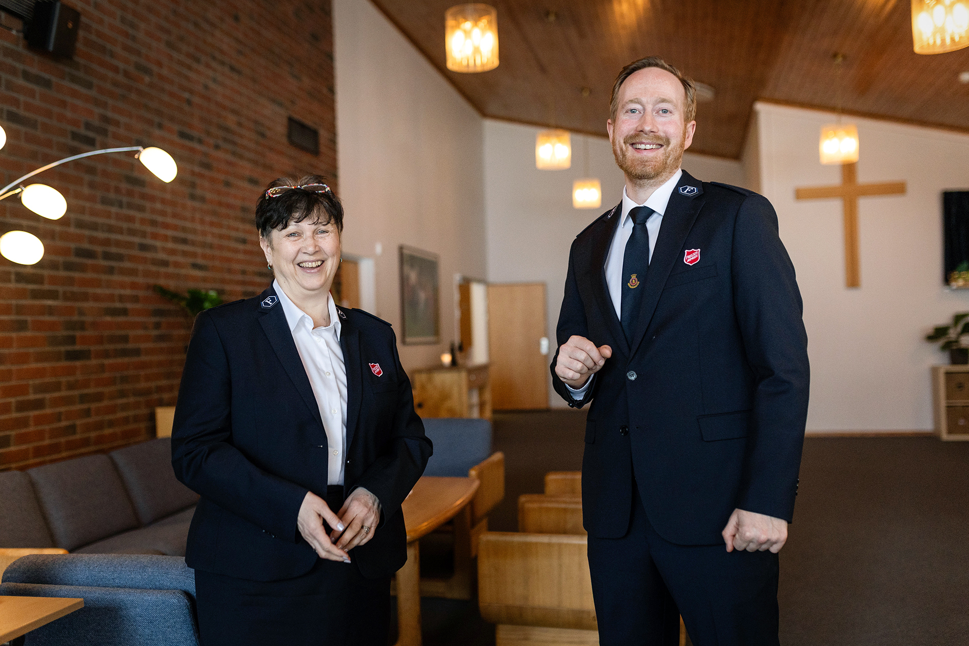 Foto: Kvinne i 60-årene med kort, mørkt hår smiler og ler sammen med en mye høyere mann i 40-årene med rødtblondt hår og skjegg.  Begge har på seg uniformer fra Frelsesarmeen og står i et kirkerom med kors i tre bak på ene veggen..