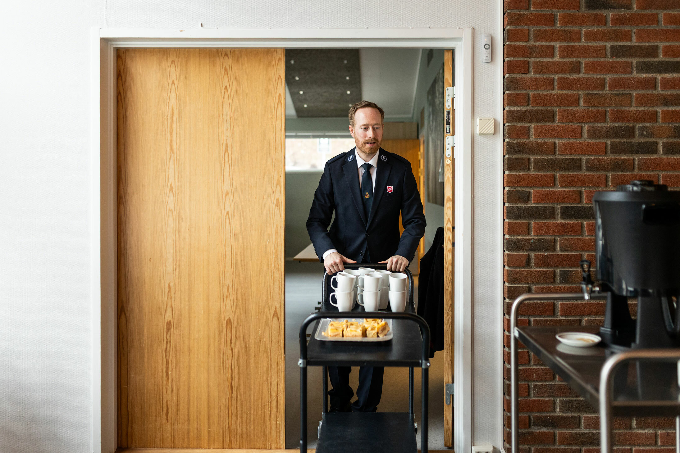 Foto: To bilder etter hverandre der en mann i 40-årene med rødblondt hår og skjegg iført uniform fra Frelsesarmeen. Han setter kaffekopper på en tralle på et stort kjøkken. På neste bilde triller han samme tralle gjennom en tredør inn til et rom med vegger av rød teglstein.