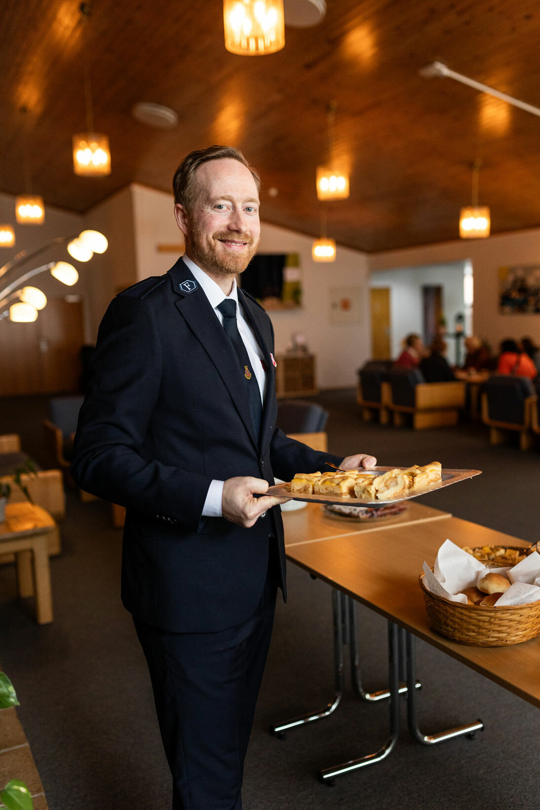 Foto: Mann i 40-årene med rødblondt hår og skjegg. Har på seg uniform fra Frelsesarmeen og står ved et bord med bakst.