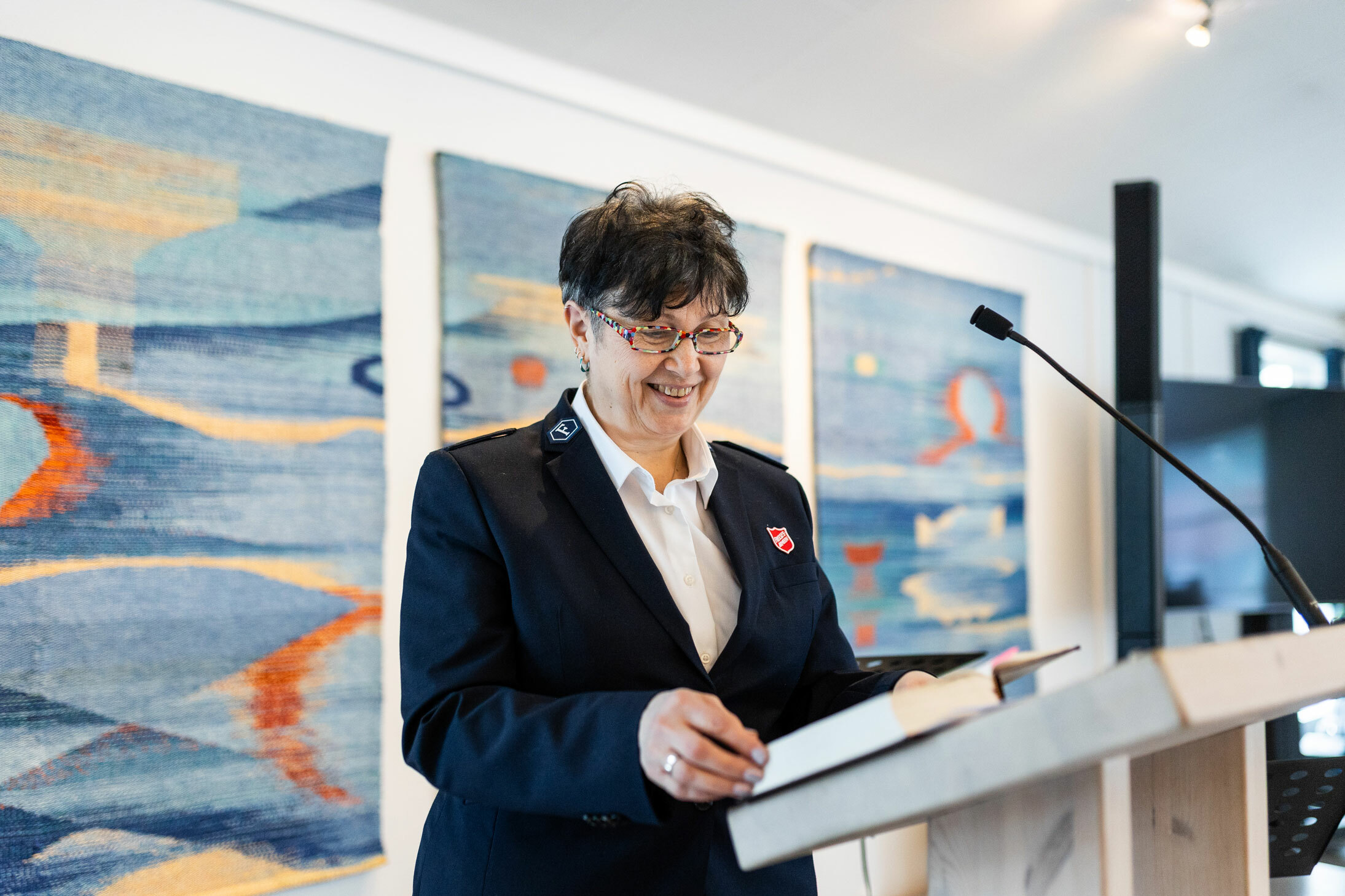 Foto: Kvinne i 60-årene med briller leser en tekst fra talerstolen i et kirkerom med vevde tepper i bakgrunnen. Hun ser ned i en bok og har på seg uniform fra Frelsesarmeen.