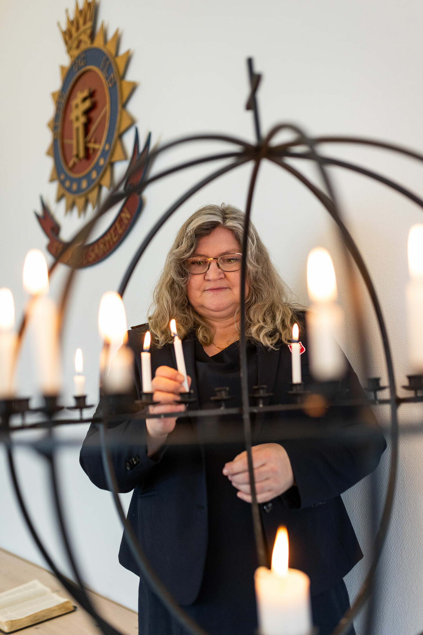 Foto: Norsk kvinne i 60-årene men halvlangt, krøllete hår og briller setter et hvitt, tent stearinlys i en svart globe med korps på toppen. I bakgrunnen sees symbolet fra Frelsesarmeen med blod og ild på veggen bak.