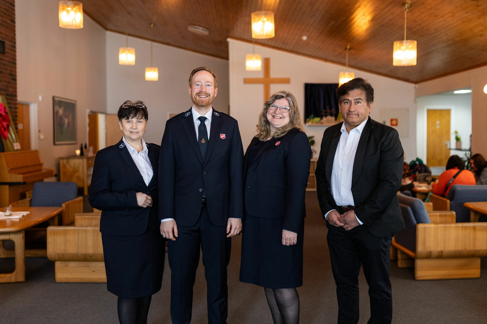 Foto: Fire personer, to kvinner og to menn, står i mørke klær i et kirkerom. Tre av dem har på seg uniformer fra Frelsesarmeen. PÅ veggen bak henger et kors i tre.