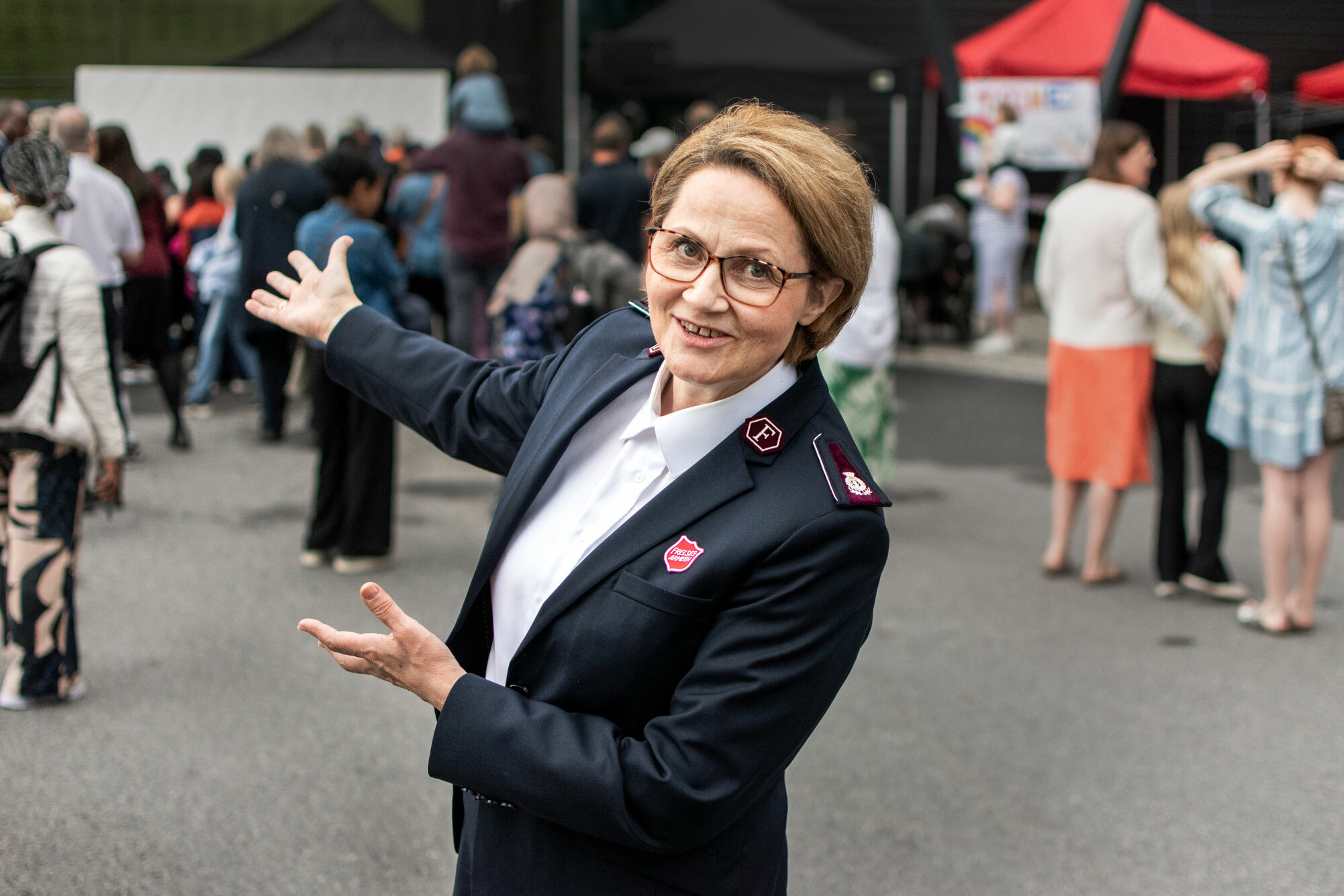 Foto: Kvinne i 60-årene med briller i uniform fra Frelsesarmeen. Hun peker innover på en plass med mennesker begge armene.