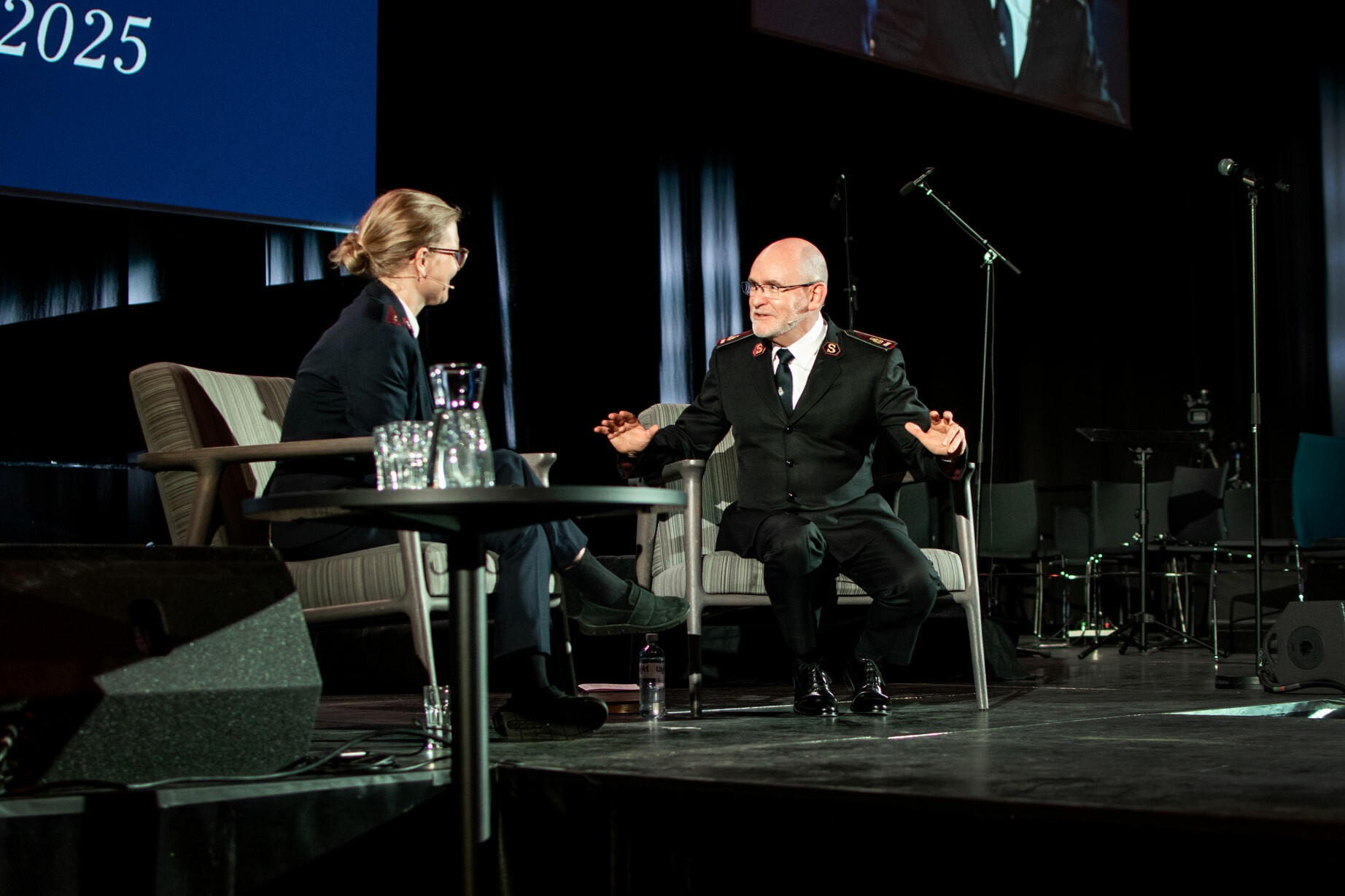 Foto: Yngre kvinne med briller og håret oppsatt sitter i en lenestol på en scene sammen med en mann i 60-årene uten hår og briller. Begge har på seg uniformer fra Frelsesarmeen.