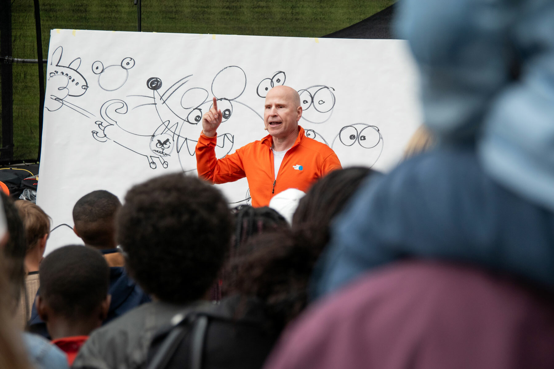 Foto: Mann uten hår i oransje fleecejakke står foran et stort, hvitt ark der han tegner figurer.