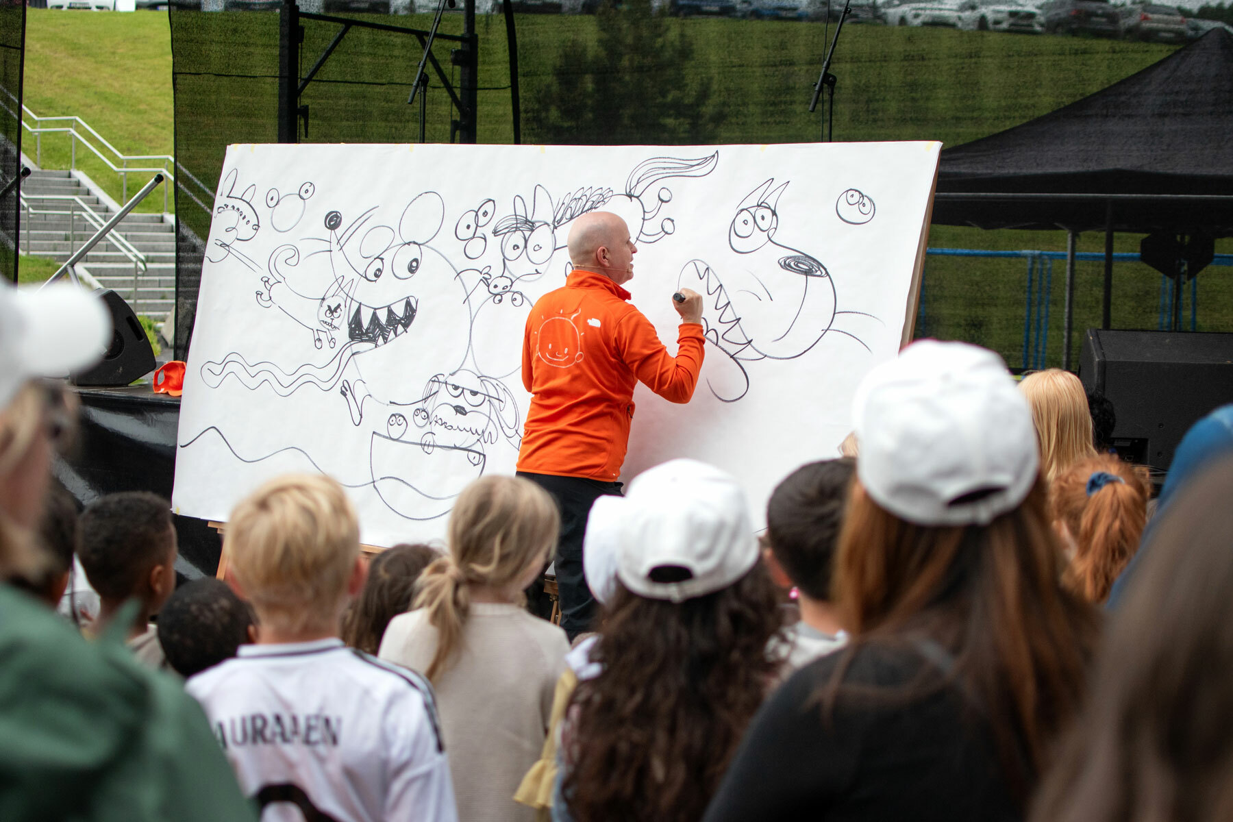 Foto: Mann uten hår i oransje fleecejakke står med ryggen til publikum og tegner på en hvit tavle.