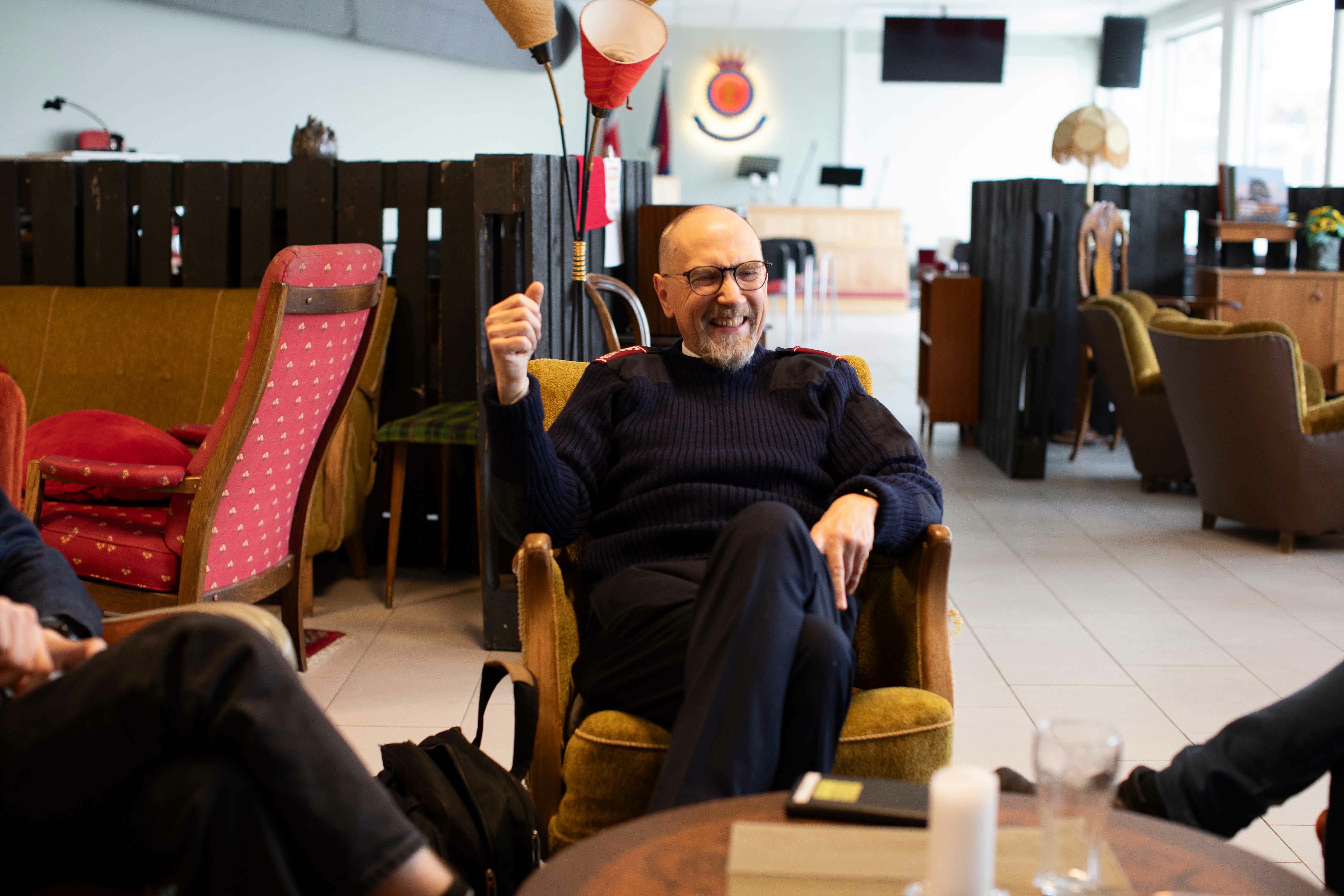 En eldre, skallet mann med marineblå ribbestrikket ullgenser med frelsesarmeens røde skulderklaffer sitter og ler og snakker med andre s. I bakgrunnen ser vi gamle møbler og et podium med Frelsesarmeens krone på veggen.