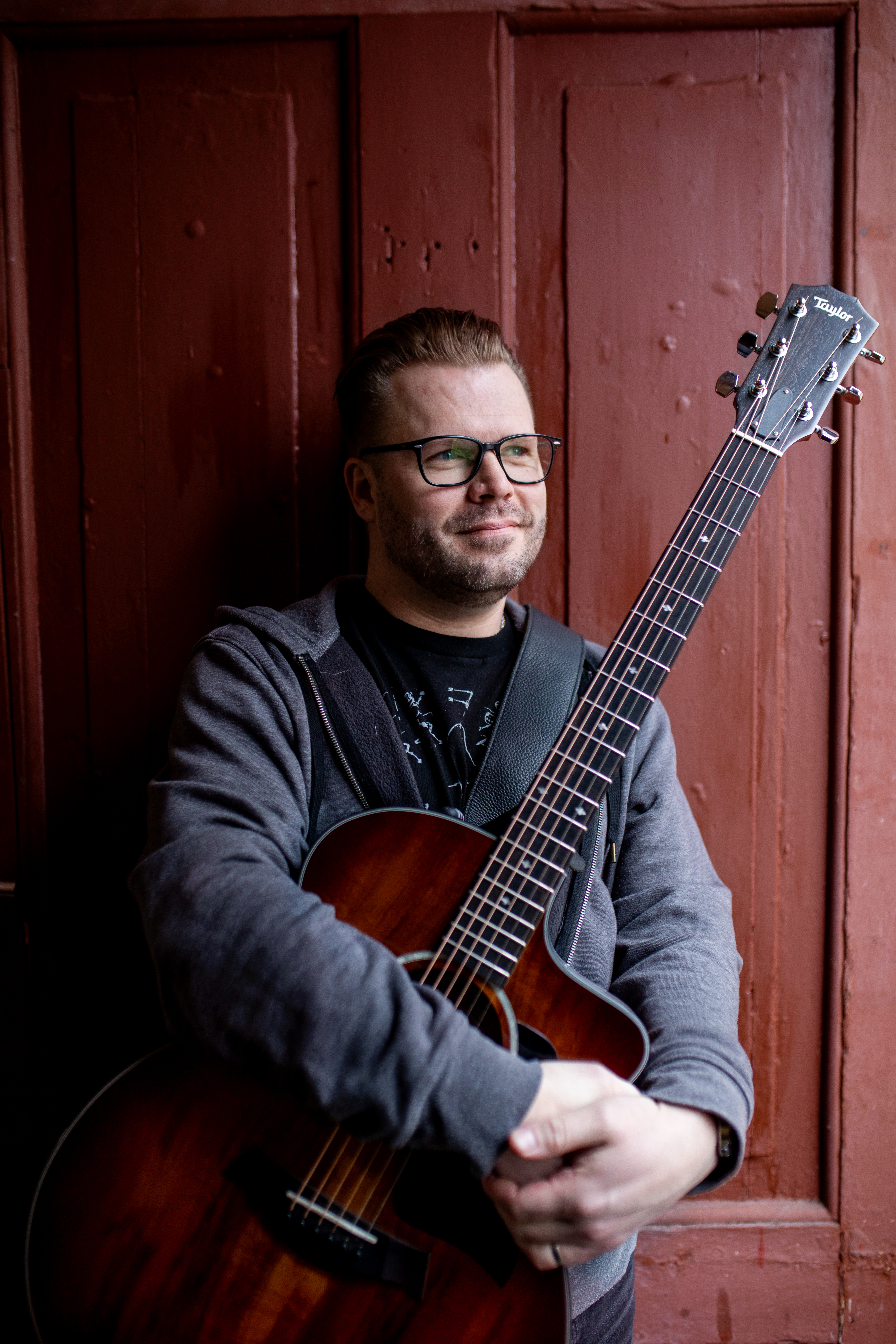 Kristian Nygård står foran en rød tredør. Han smiler og holder gitaren sitt tett inntil seg.