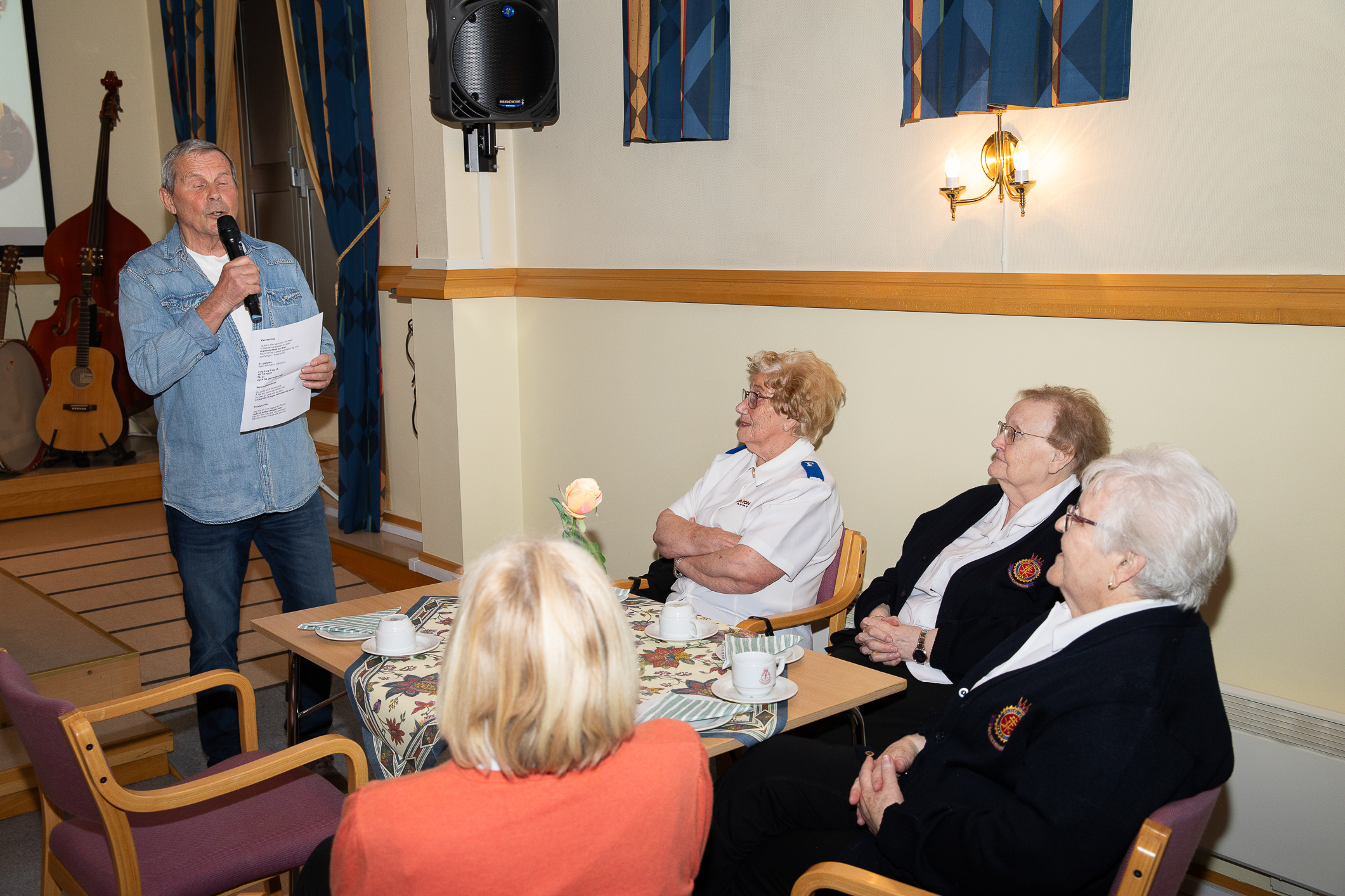 En middelaldrende mann synger  på et møte i Frelsesarmeen. Fire voksne kvinner sitter rundt et bord med duk og hører på. Flere har frelsesarmeuniform.