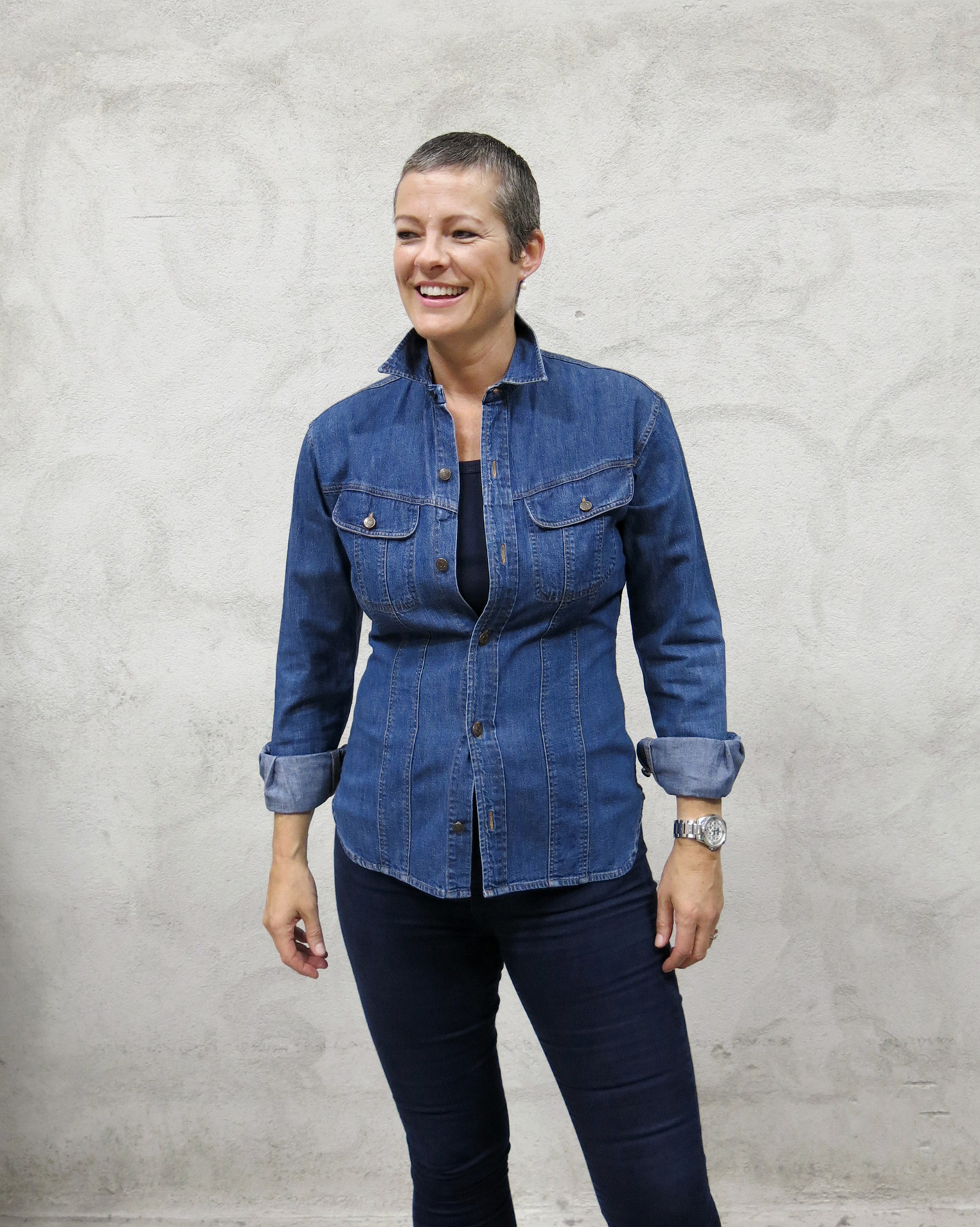 Kristin Hareide med kort sveis og blå denim skjorte, smilende foran en grå murvegg