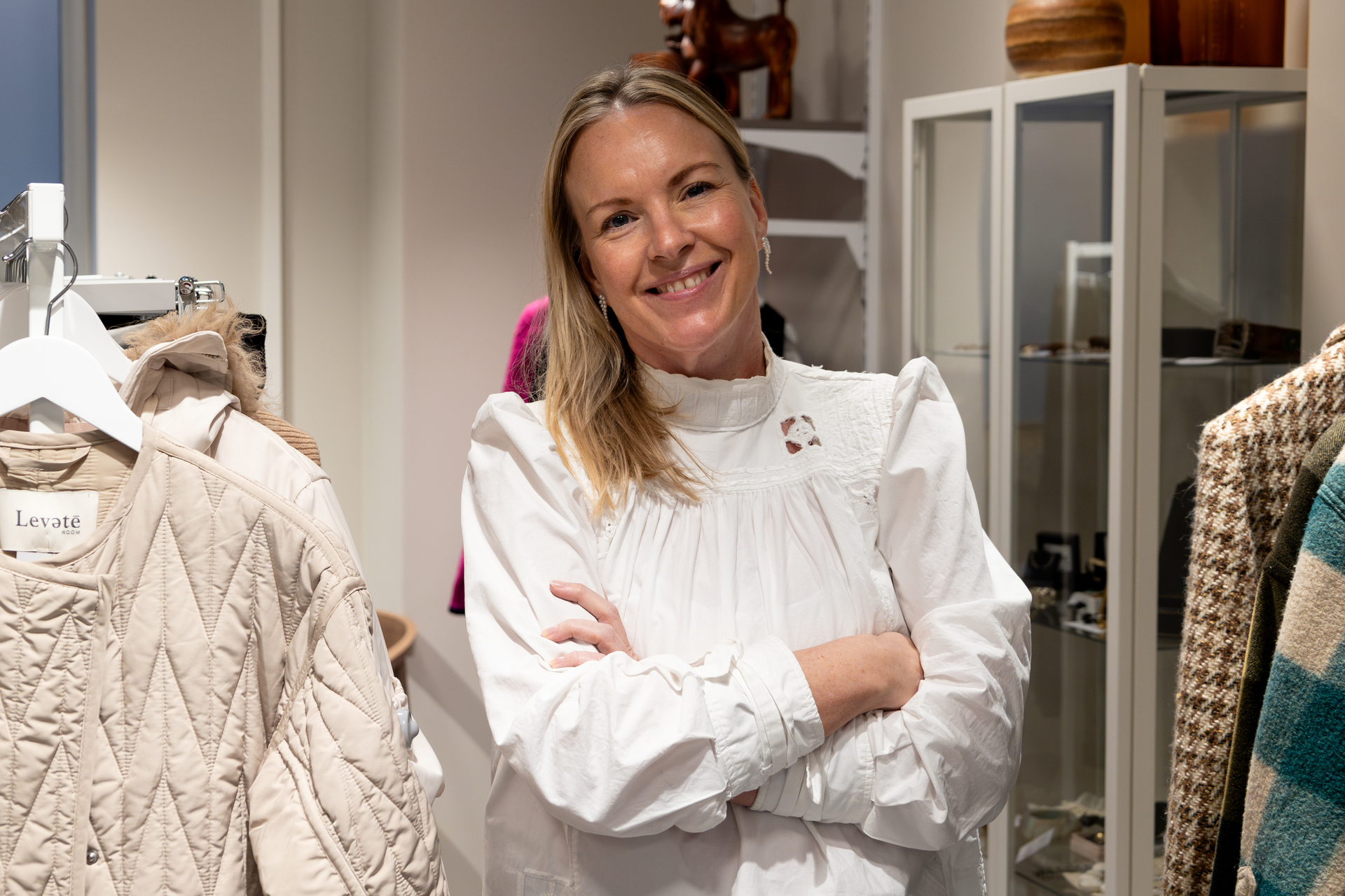 Foto: Kvinne i 50-årene i hvite bluse og langt, blondt hår smiler i en butikk med pene, brukte klær på stativ og h