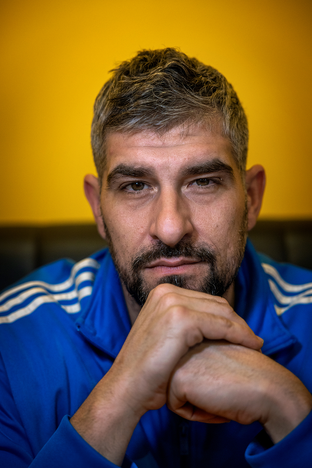Foto: Mann med grålig hår og skjegg stirrer intenst i kamera. Veggen bal er knall gul.