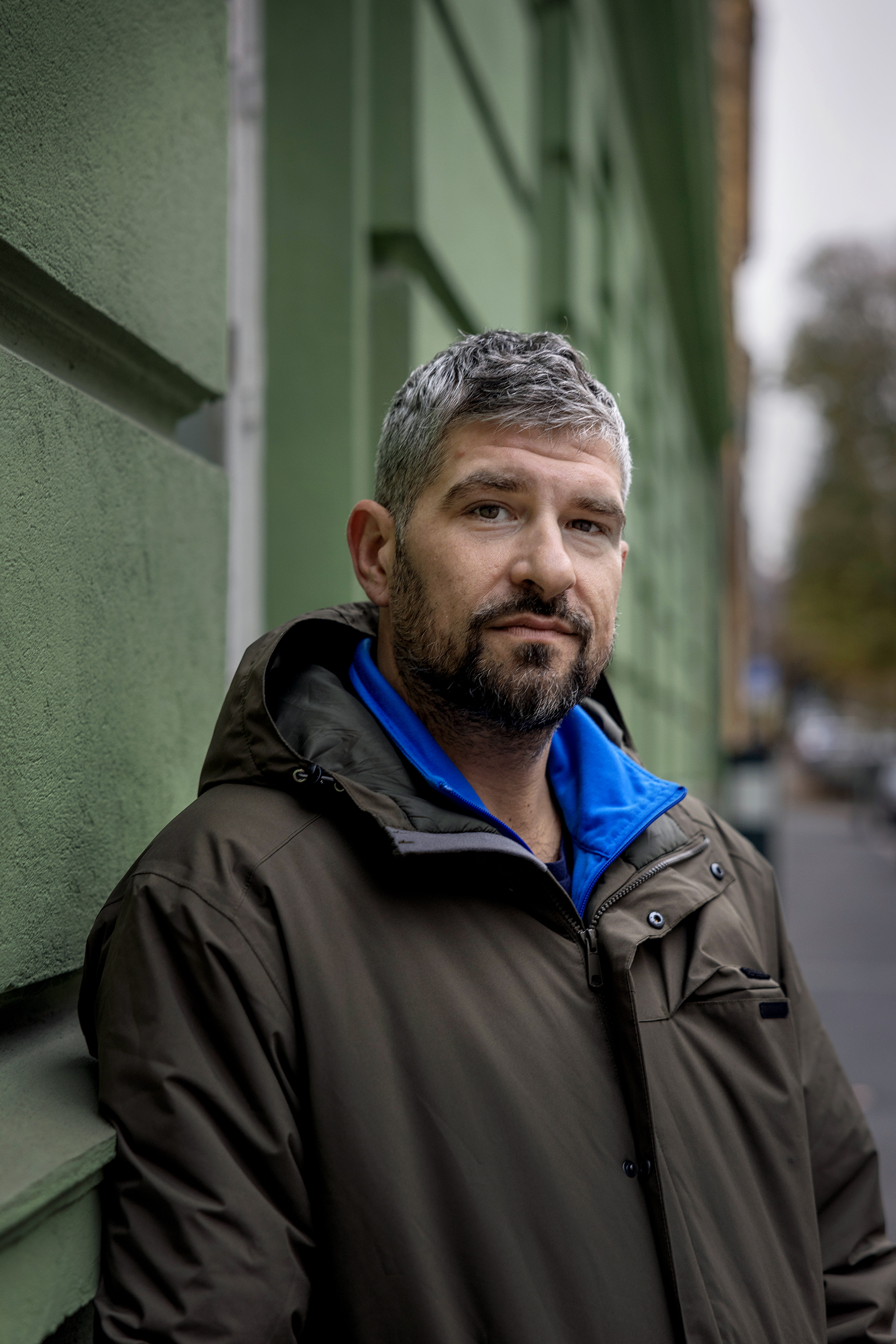 Foto: Mann med grålig hår og skjegg står ute i bybildet, foran en grønn murvegg.