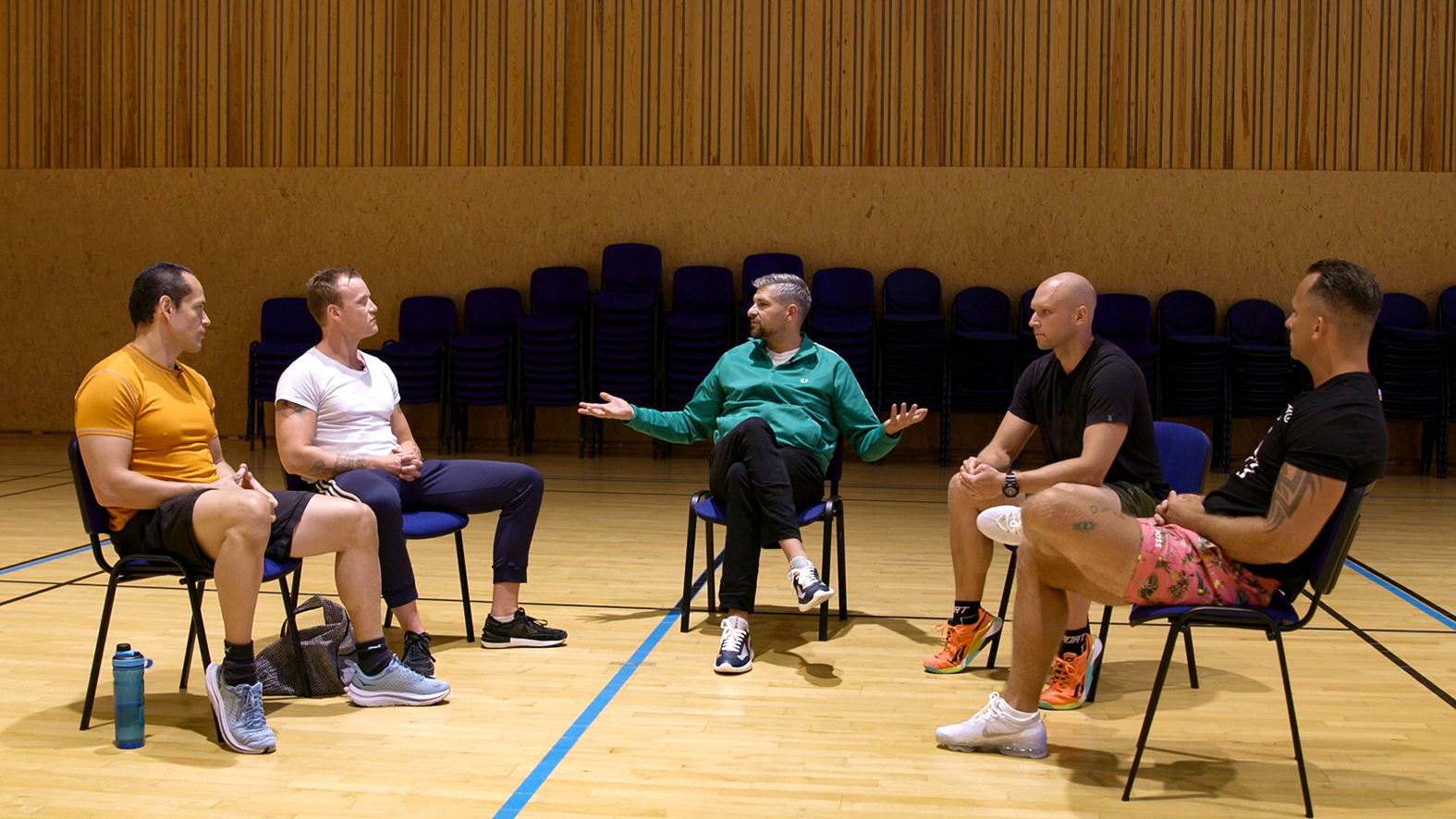 Foto: Fem menn sitter på hver sin stol i en halvsirkel inne i en gymsal. Alle er iført treningstøy. Mannen i midten har slått ut armene.