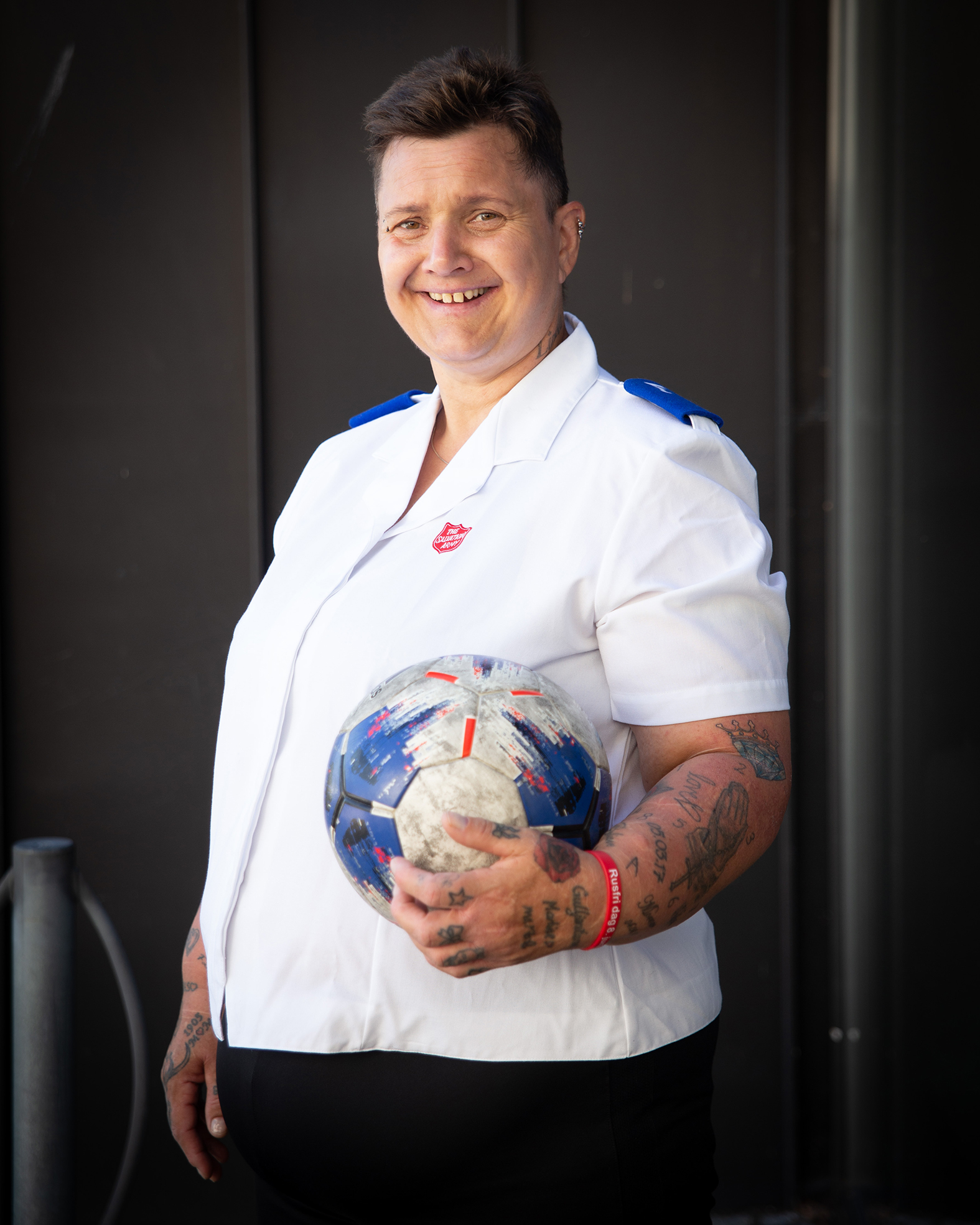 Bildet viser Linda som står foran en mørk vegg. Hun har kort hår og Frelsesarme-t-skjorte og holder en fotball.