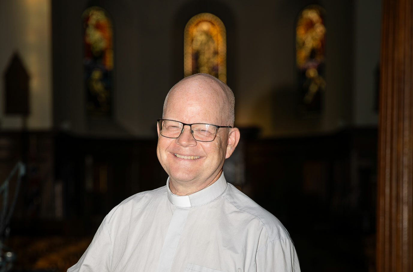 Prest står i mørkt kirkerom. Kan se glassmalerier i bakgrunnen.