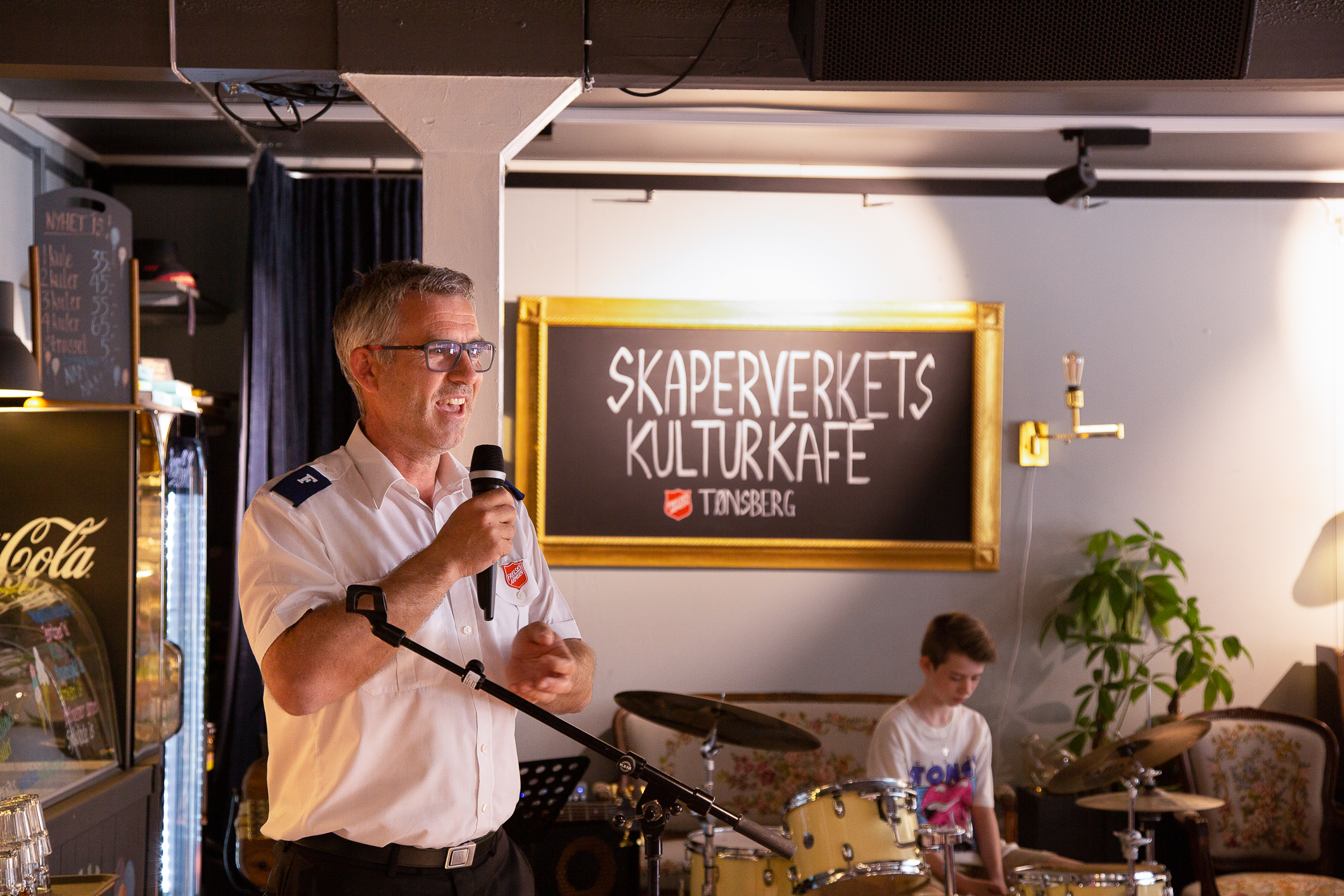 Mann i uniform taler fra hjørnet av kafeen. På den store tavla i bakgrunnen står: Skaperverket kafe Tønsberg.
