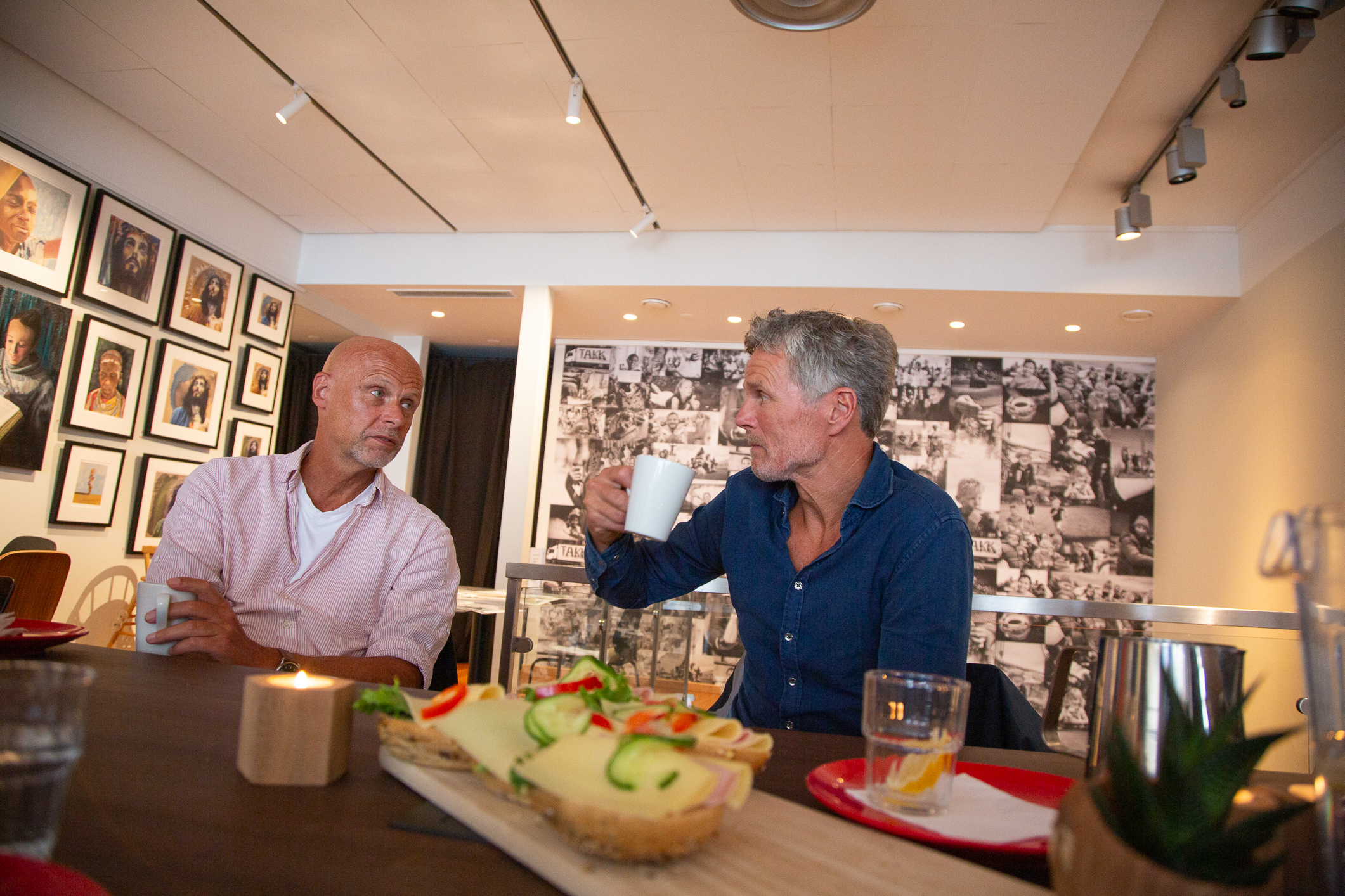 To menn sitter ved et bord, drikker kaffe og prater med hverandre. Foran dem står et brett med påsmurte rundstykker.