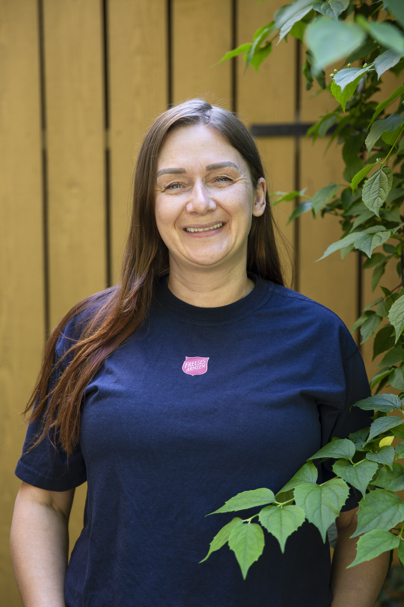 Foto: Kvinne med halvlangt, mørkt hår står ute foran en gulmalt vegg. Hun har på seg blå T-skjorte med rød logo fra Frelsesarmeen.