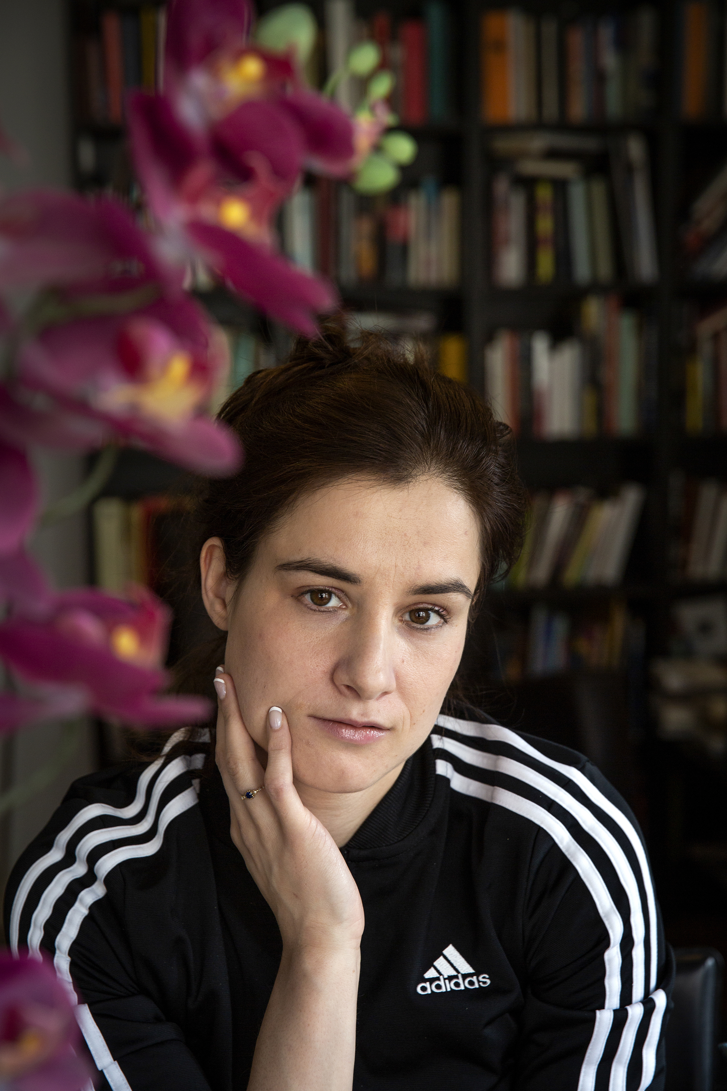 Maria Kjos Fonn har på seg Adidas trengingsdress, i bakgrunnen er det en full bokhylle og i forgrunnen en lilla orkidè blomst.