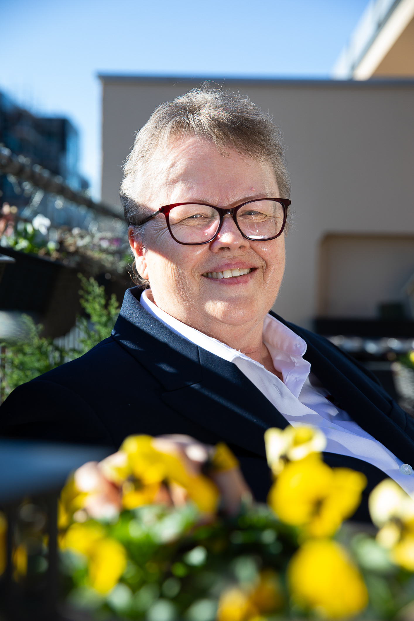 Eldre kvinne med briller og blå uniformsjakke med Frelsesarmeens røde skjold på jakken sitter i stuen, sitter og smiler på balkongen bak gule blomster.
