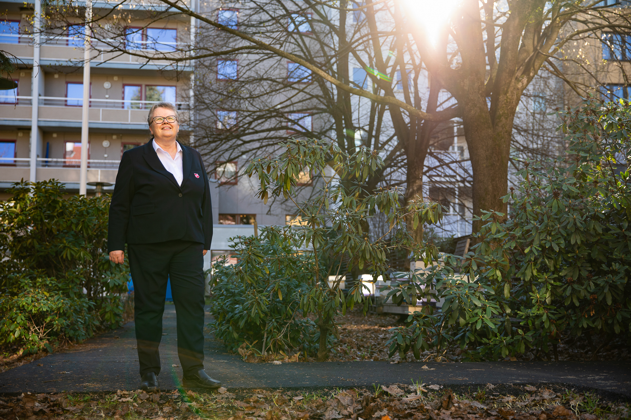 Eldre kvinne med briller og blå jakke med Frelsesarmeens røde skjold på, står og ser oppover. Bak henne er det grå blokker, busker og trær.