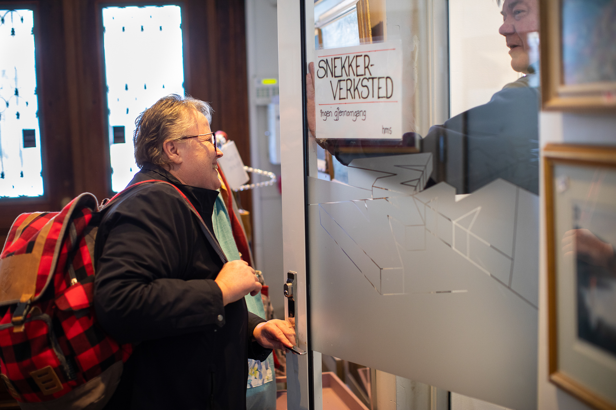 En eldre kvinne med blå jakke og rødrutete ryggsekk åpner en glassdør som det står «Snekkerverksted» på. En mann innenfor hjelper henne å åpne døren.