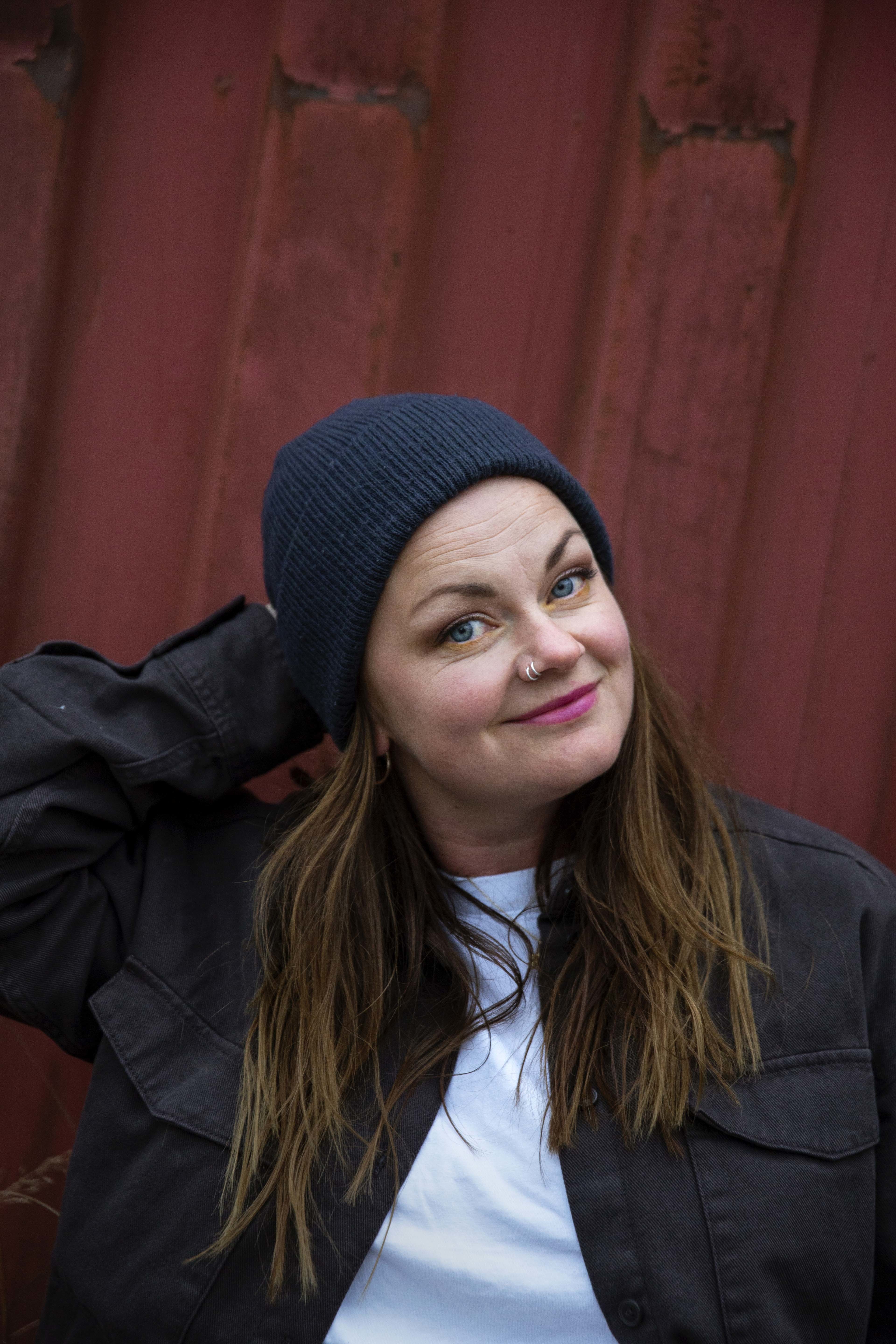 Marianne Sveen med lue og brunt hår, foran en rød husvegg
