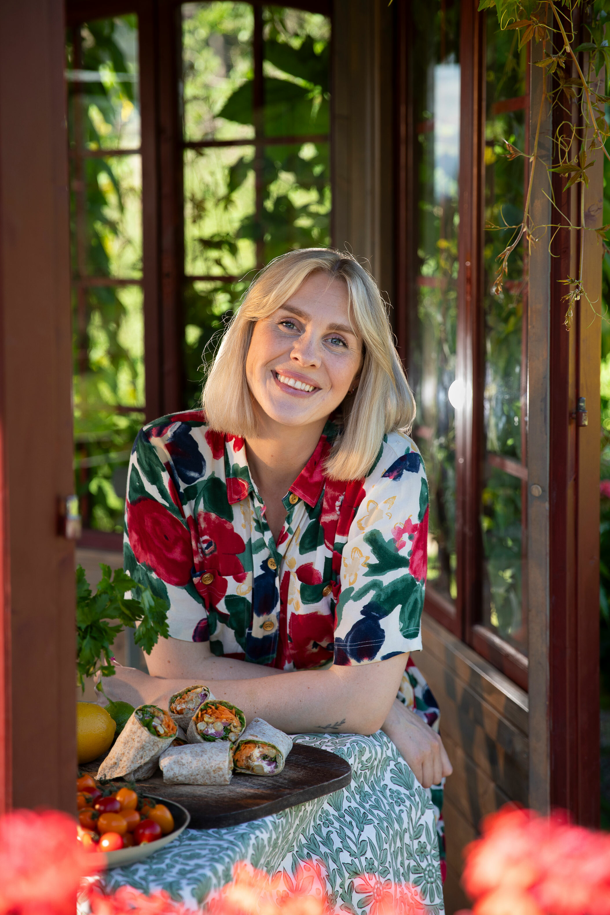 Foto: Ung kvinne i blomstrete kjole sitter ute i en vinterhage. På bordet foran seg ligger en tallerken med flere wraps.