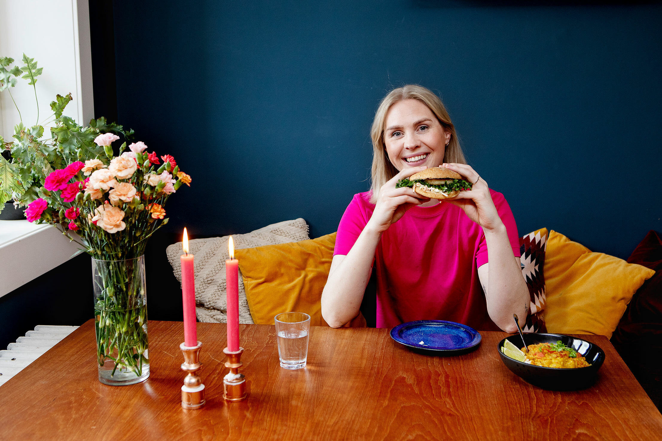 Foto: Ung kvinne med halvlangt, blondt hår sitter ved et kjøkkensbord pyntet med blomster og stearinlys. Hun holder opp en burger mellom hendene og smiler.