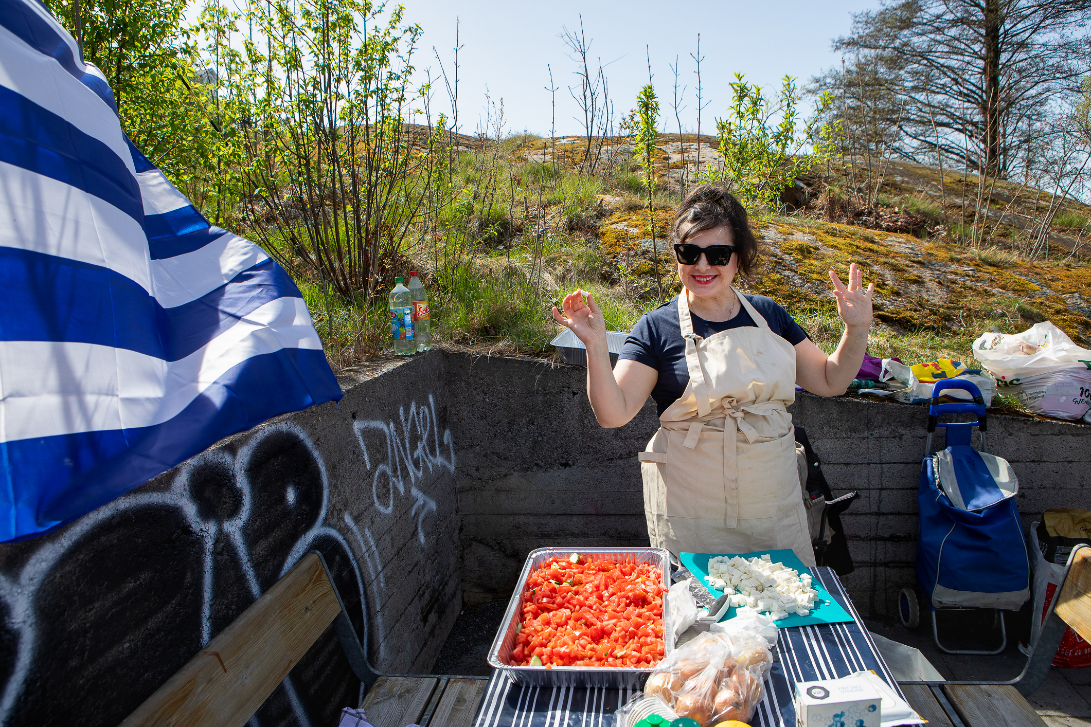En kvinne med solbriller og forkle forbereder grillmat på et utebod. Gresk flagg vaier i bakgrunnen.