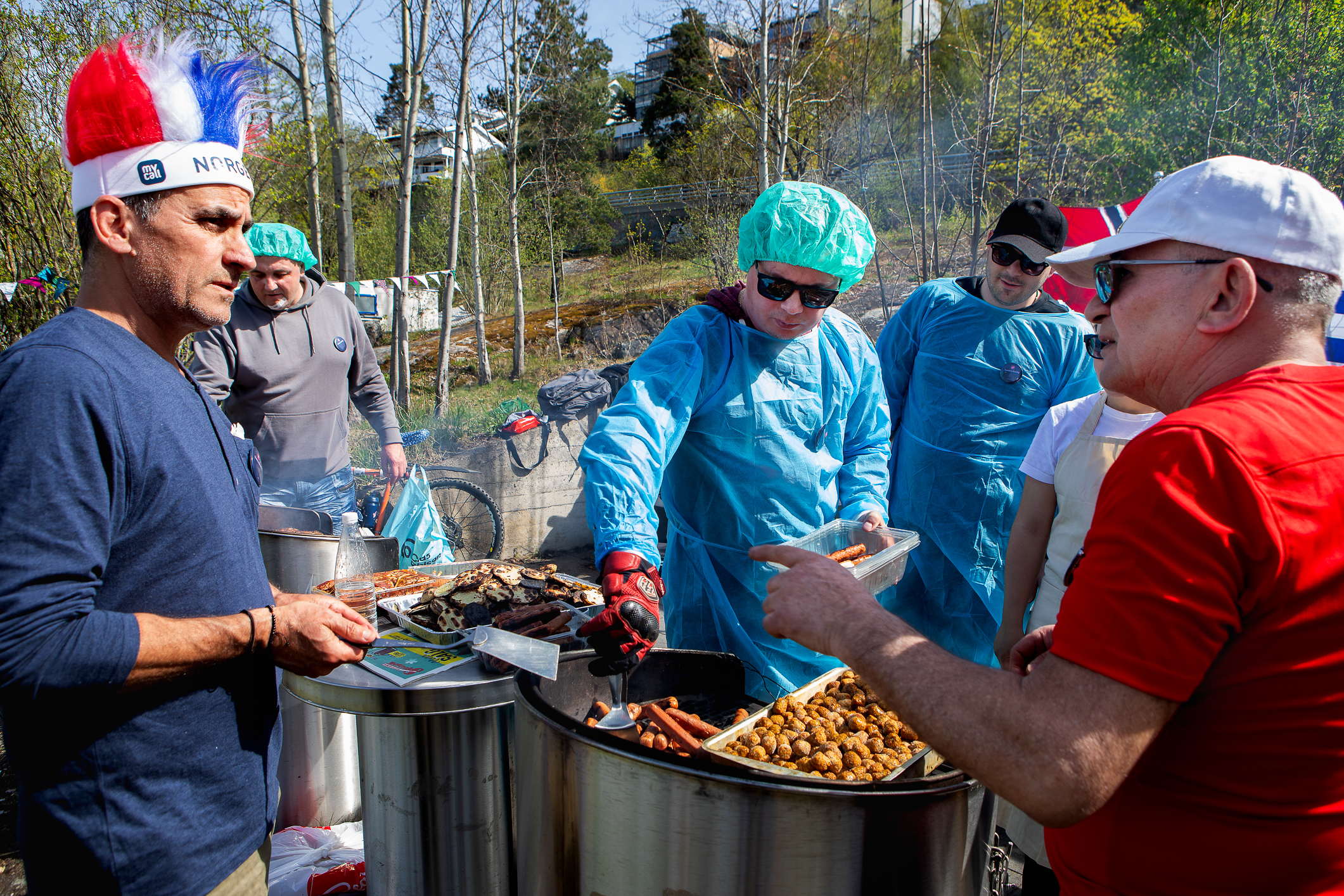 Grilling av kjøtt utendørs. Flere menn står rundt grillene. De som jobber med maten har turkise plasthetter og -frakker