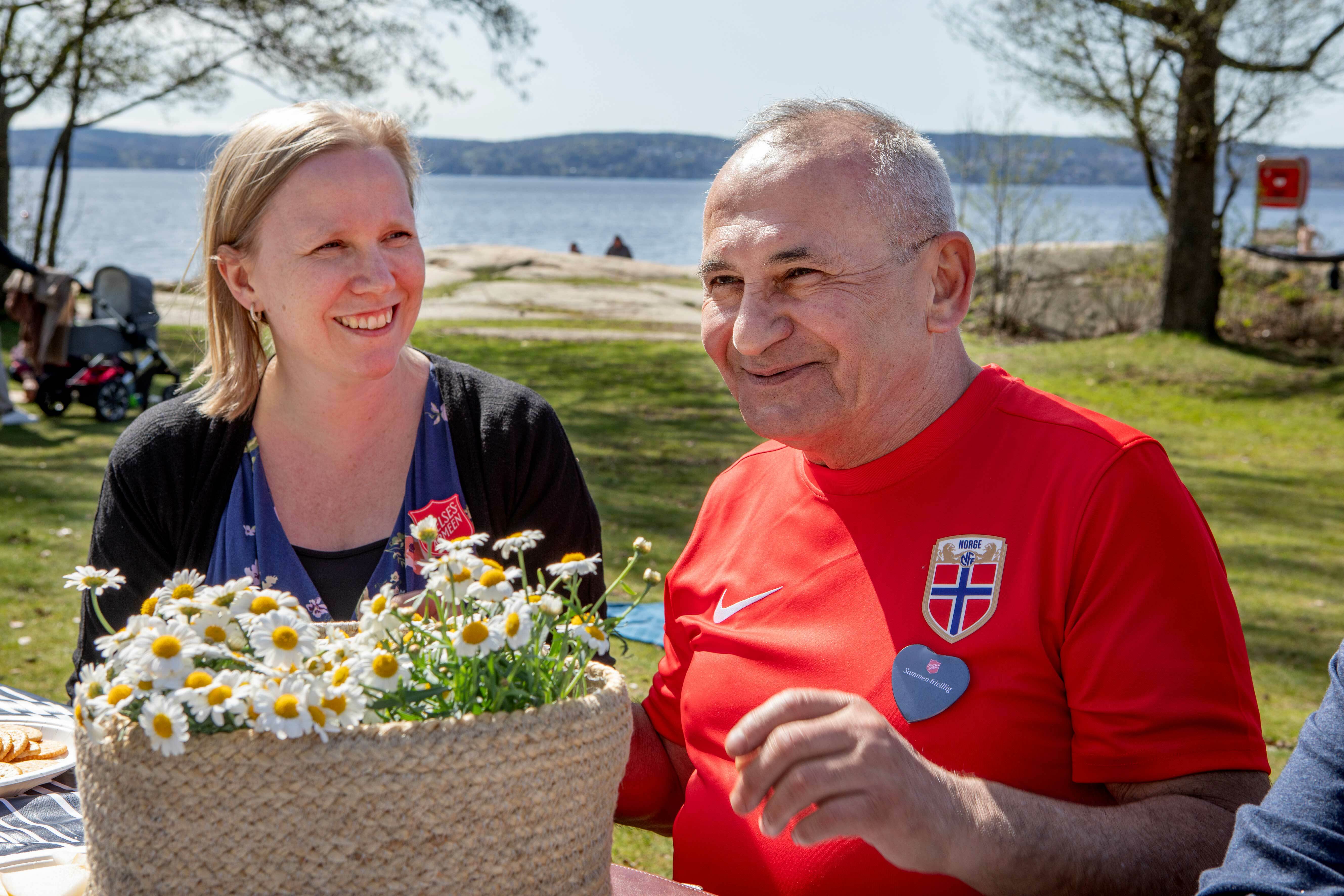 En ung kvinne og en eldre mann sitter ved et utebord på stranden og smiler mens de spiser. De har en kurv med hvite prestekrager foran seg.