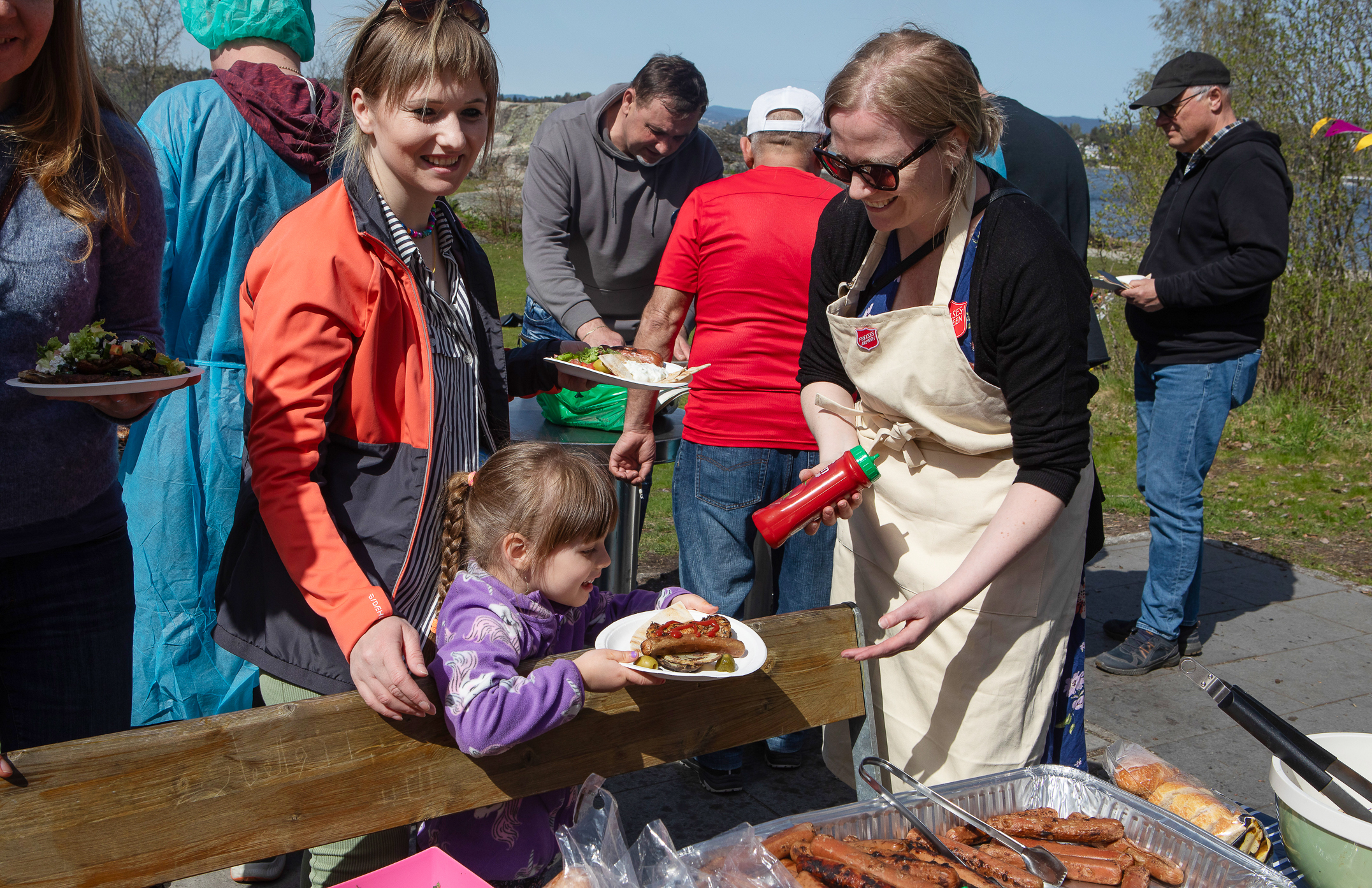 En mor og et barn forsyner seg med grillmat ute i naturen. Rundt er det mange mennesker og en kvinne med Frelsesarmeens skjold på forkleet tilbyr ketchup og en hjelpende hånd.
