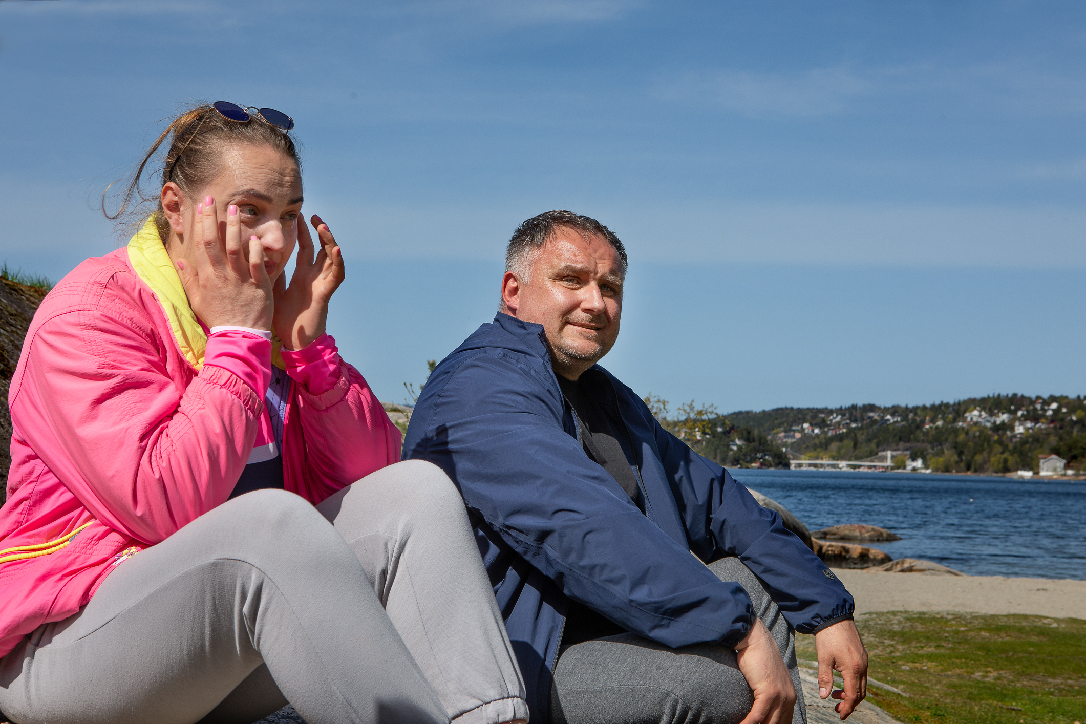 En kvinne og en mann sitter på et svaberg nede ved stranden og forteller. Hun har en rosa jakke, han har blå.