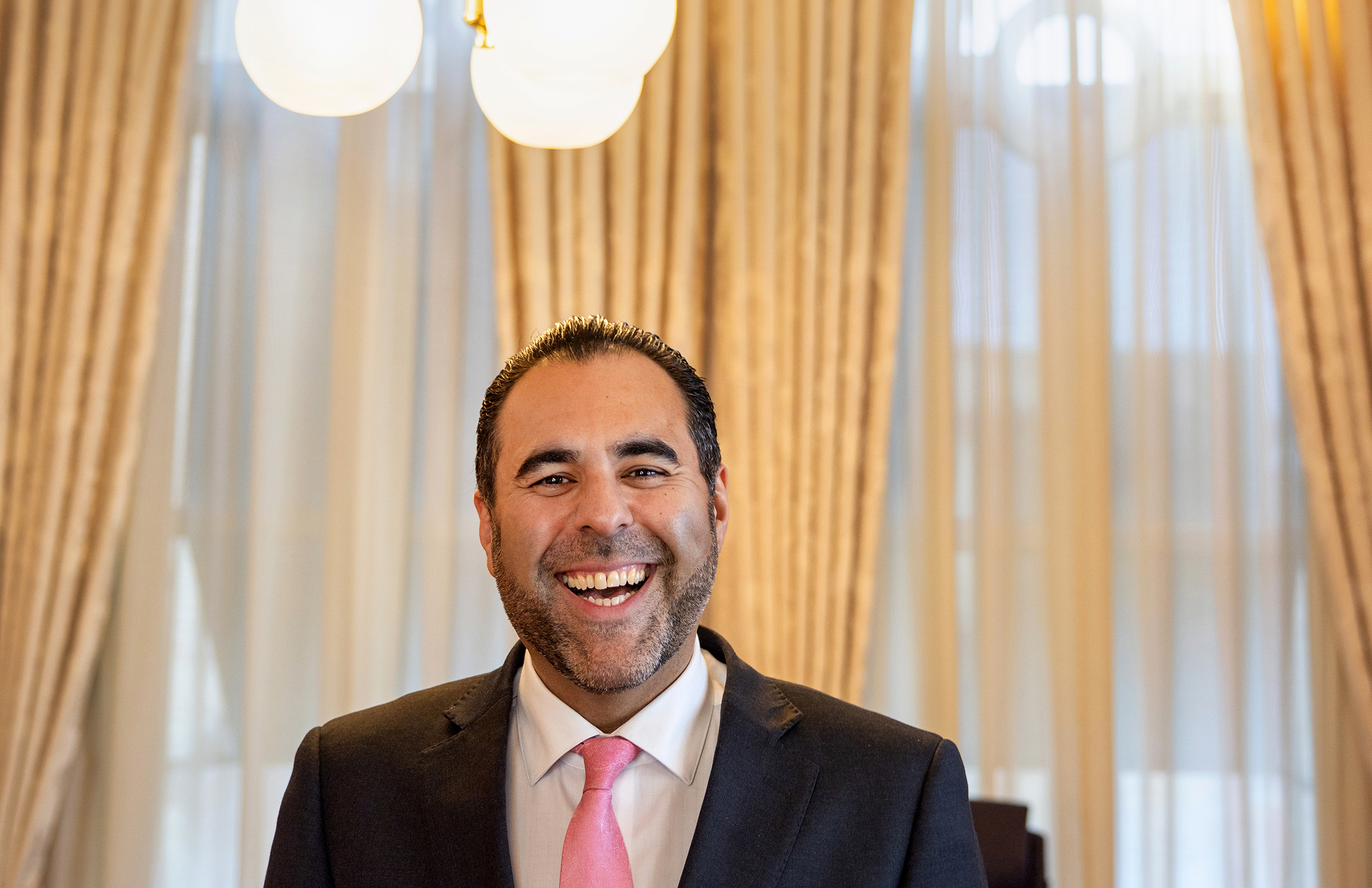 Foto: Mann i svart dress, hvit skjorte og rosa slips smiler og ler inne på kontoret sitt på Stortinget.