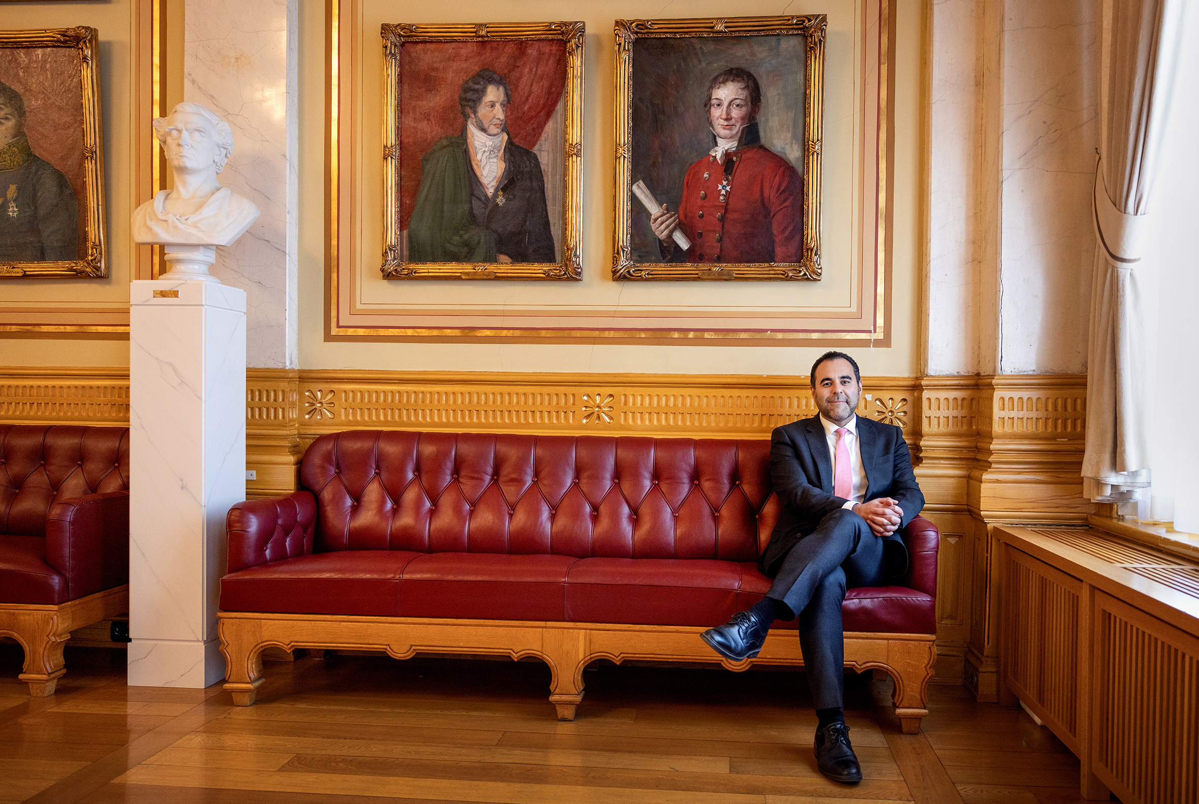 Foro: Mann som sitter på en rød, gammel skinnsofa i et historisk rom med gamle portretter på veggene.