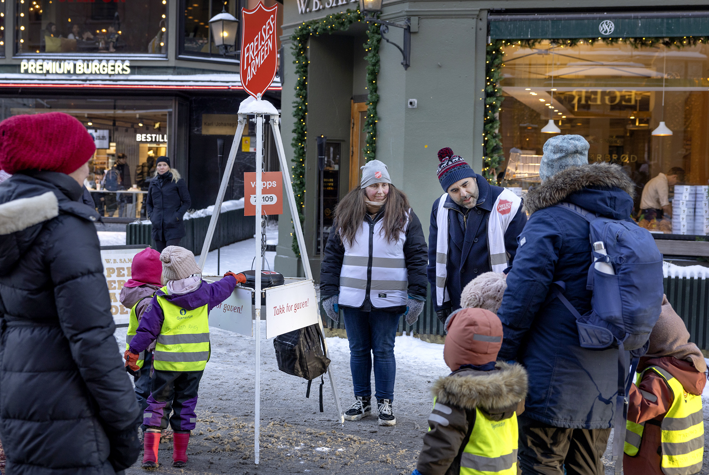 Foto: Fem barnehagebarn i gule refleksvester står sammen med voksne. De skal gi penger i Frelsesarmeens julegryte som henger på et stativ med logoen til Frelsesarmeen på toppen. Der står ei dame fra Frelsesarmeen og en mann med mørkt hår og skjegg fra Iran. Begge disse har på seg hvite vester fra Frelsesarmeen.