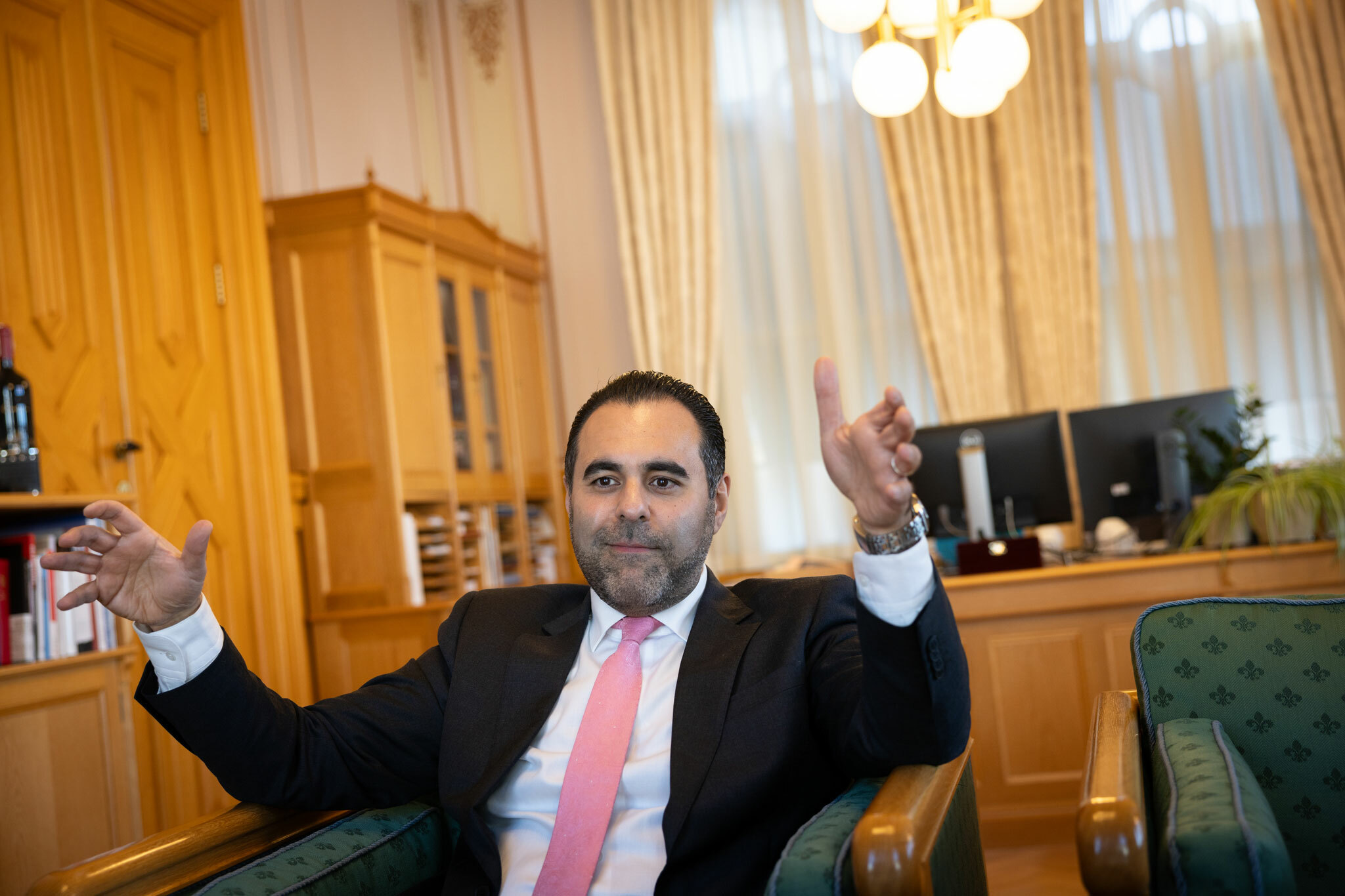 Foto: Mann i svart dress, hvit skjorte og rosa slips sitter i en lenestol inne på et stort kontor på Stortinget. Han holder opp begge armene mens han snakker.