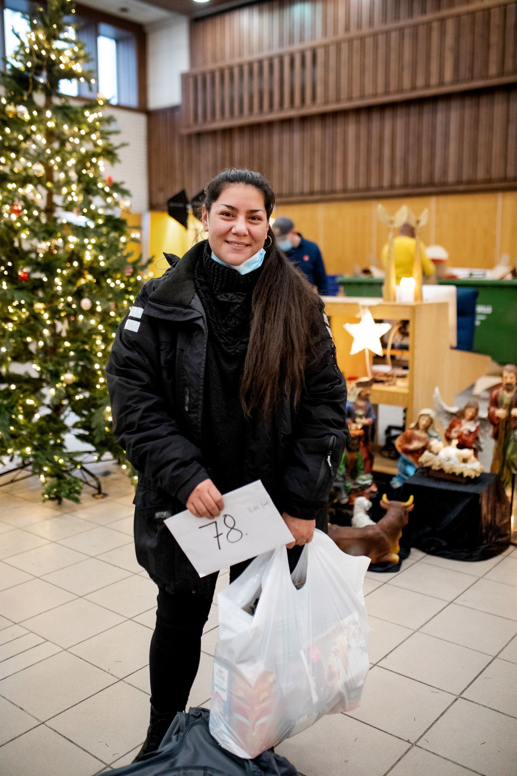 en dame står ved juletreet inne i Templet korps med en pose med mat i hånda. en mann utenfor Templet bærer en eske med mat.