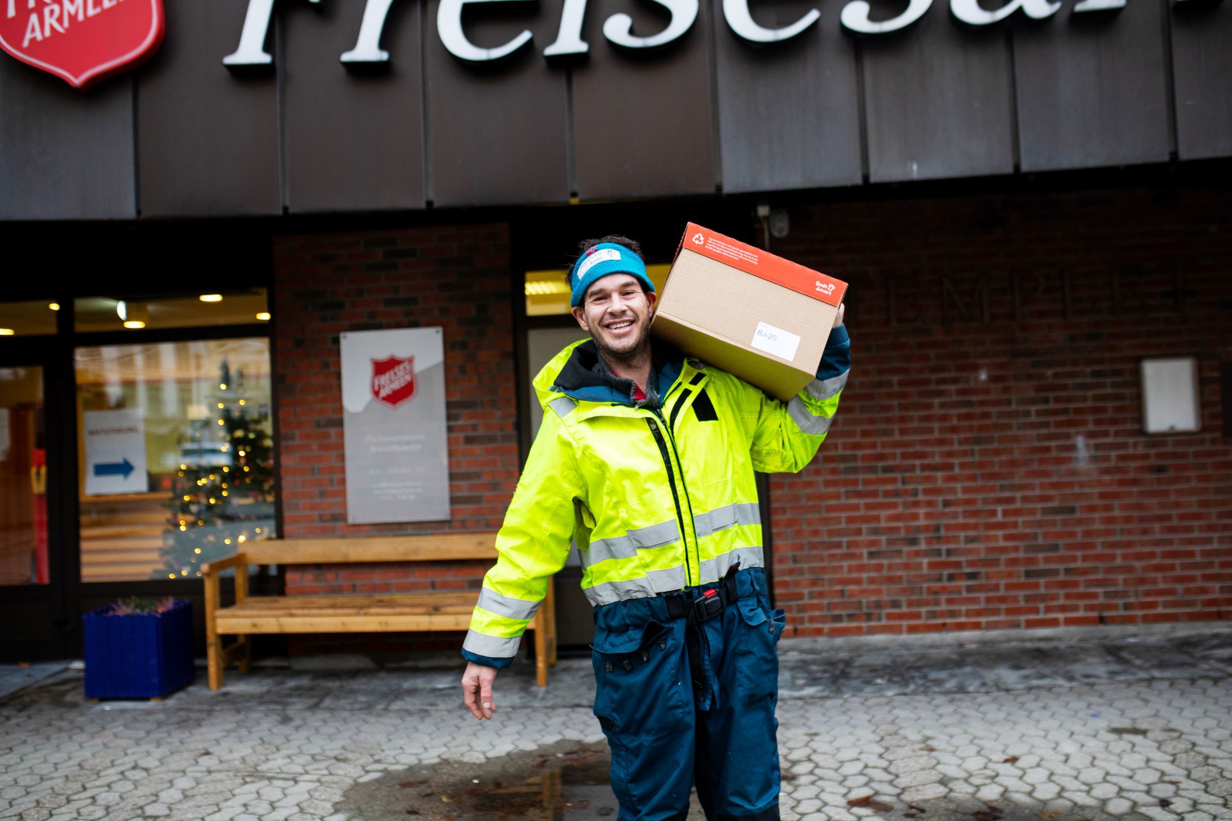 en dame står ved juletreet inne i Templet korps med en pose med mat i hånda. en mann utenfor Templet bærer en eske med mat.