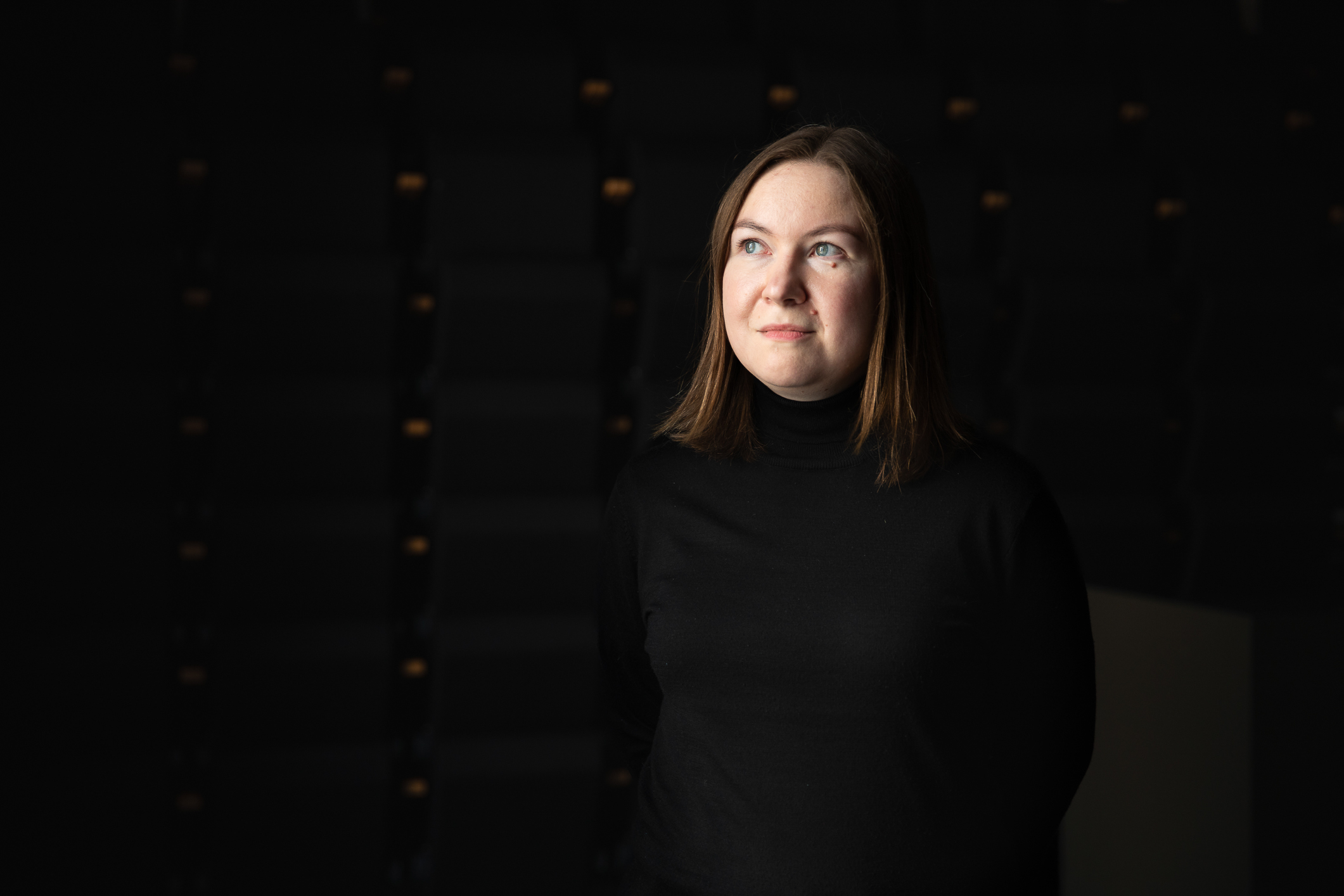 Foto: Portrettbilde av en kvinne med svarte klær i et mørkt rom. Hun har mørkeblondt, langt hår og er i slutten av 30-årene.