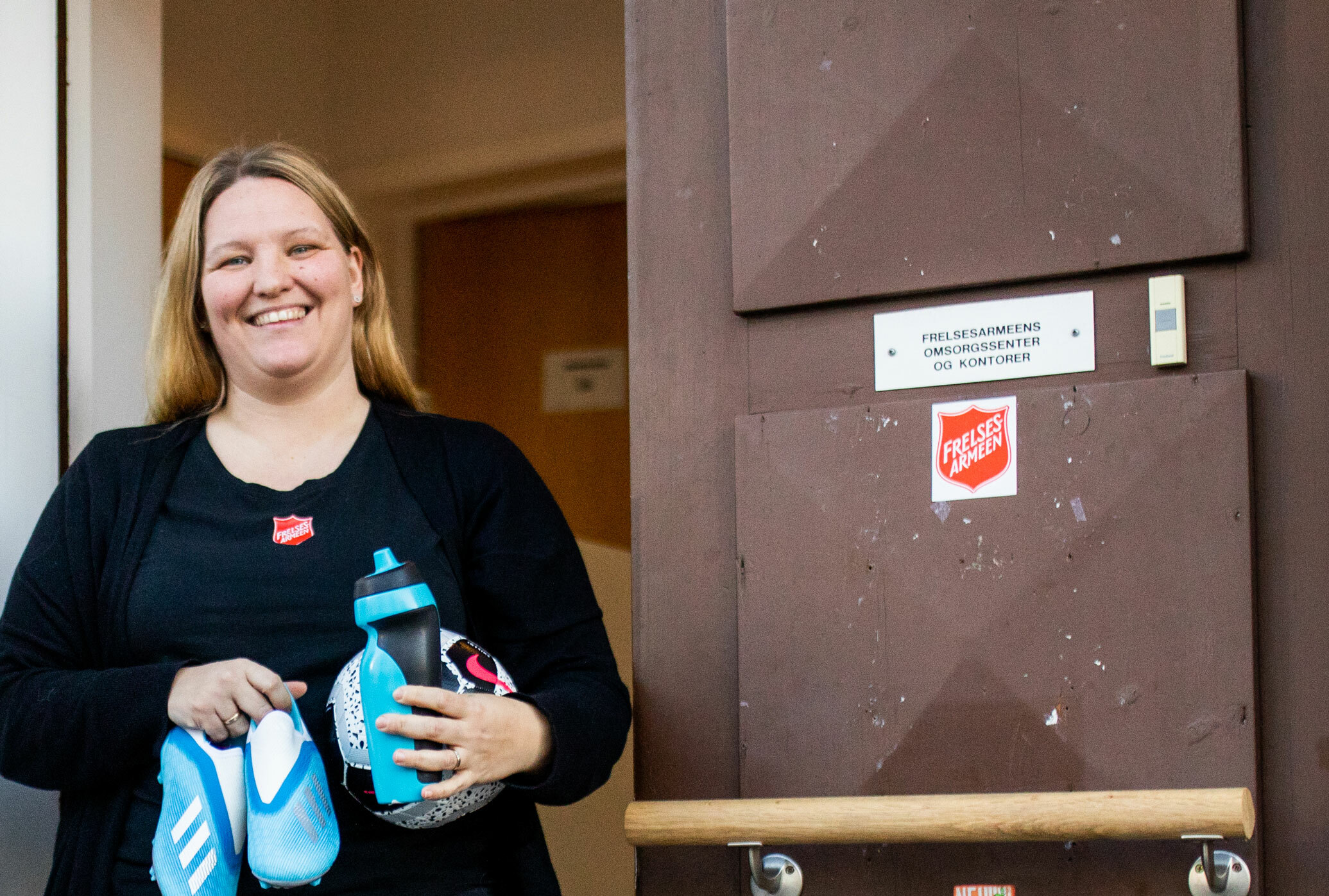 Foto: Silende kvinne står utenfor inngangsdøren til Frelsesarmeen i Drammen og holder på en vannflaske, en fotball og et par fotballsko.
